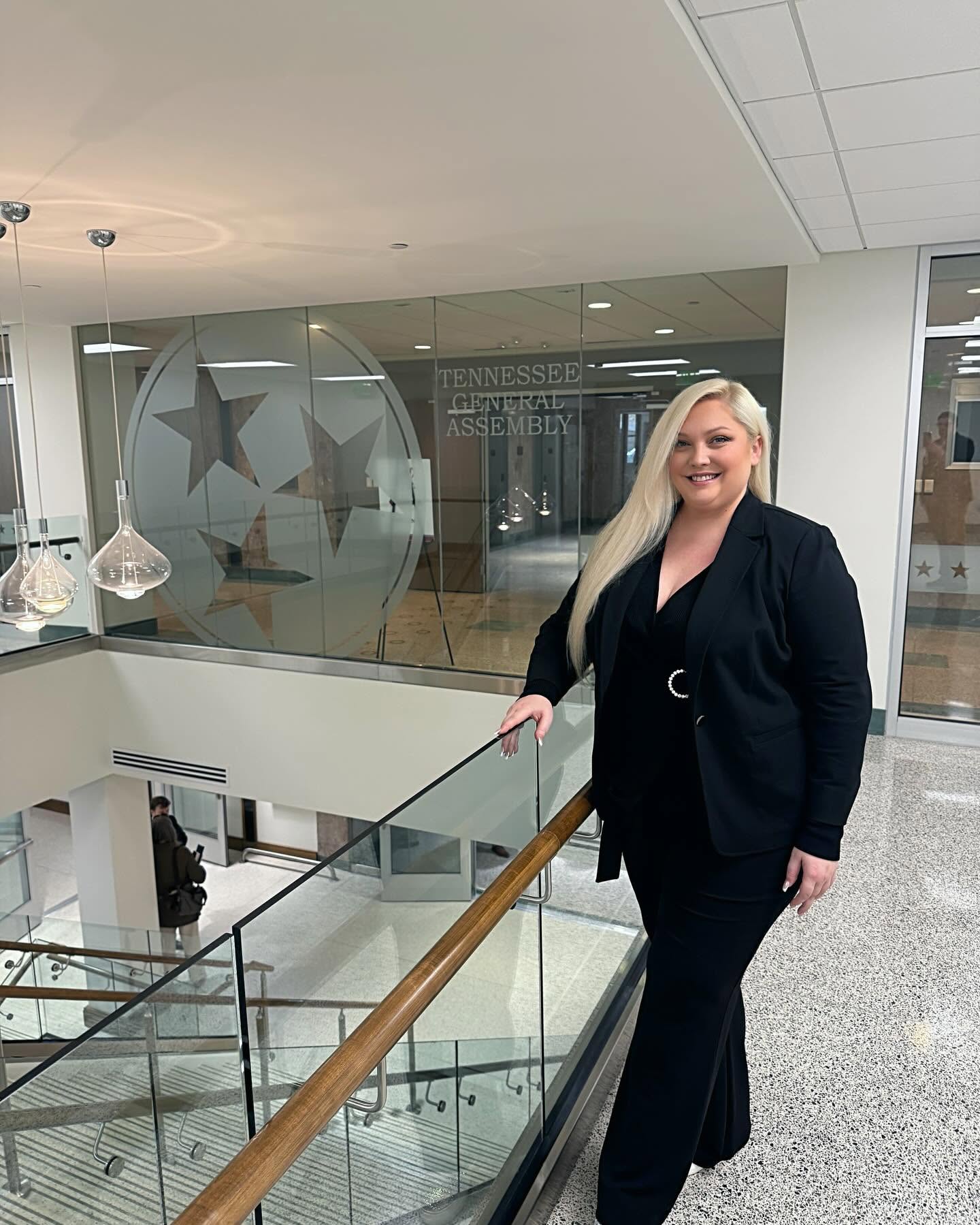 Woman with long blonde hair in a black suit standing by glass railing inside building, smiling, with star decorations on glass wall behind.
