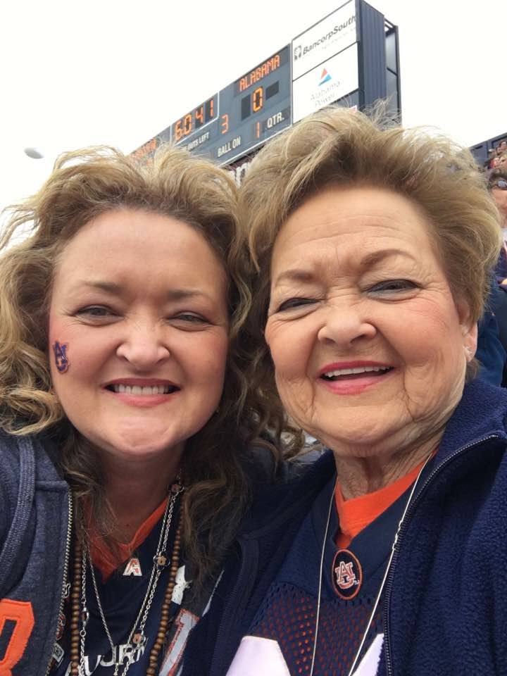 Two women smiling outdoors with a scoreboard in the background, close-up selfie, one with curly hair and the other with short hair.