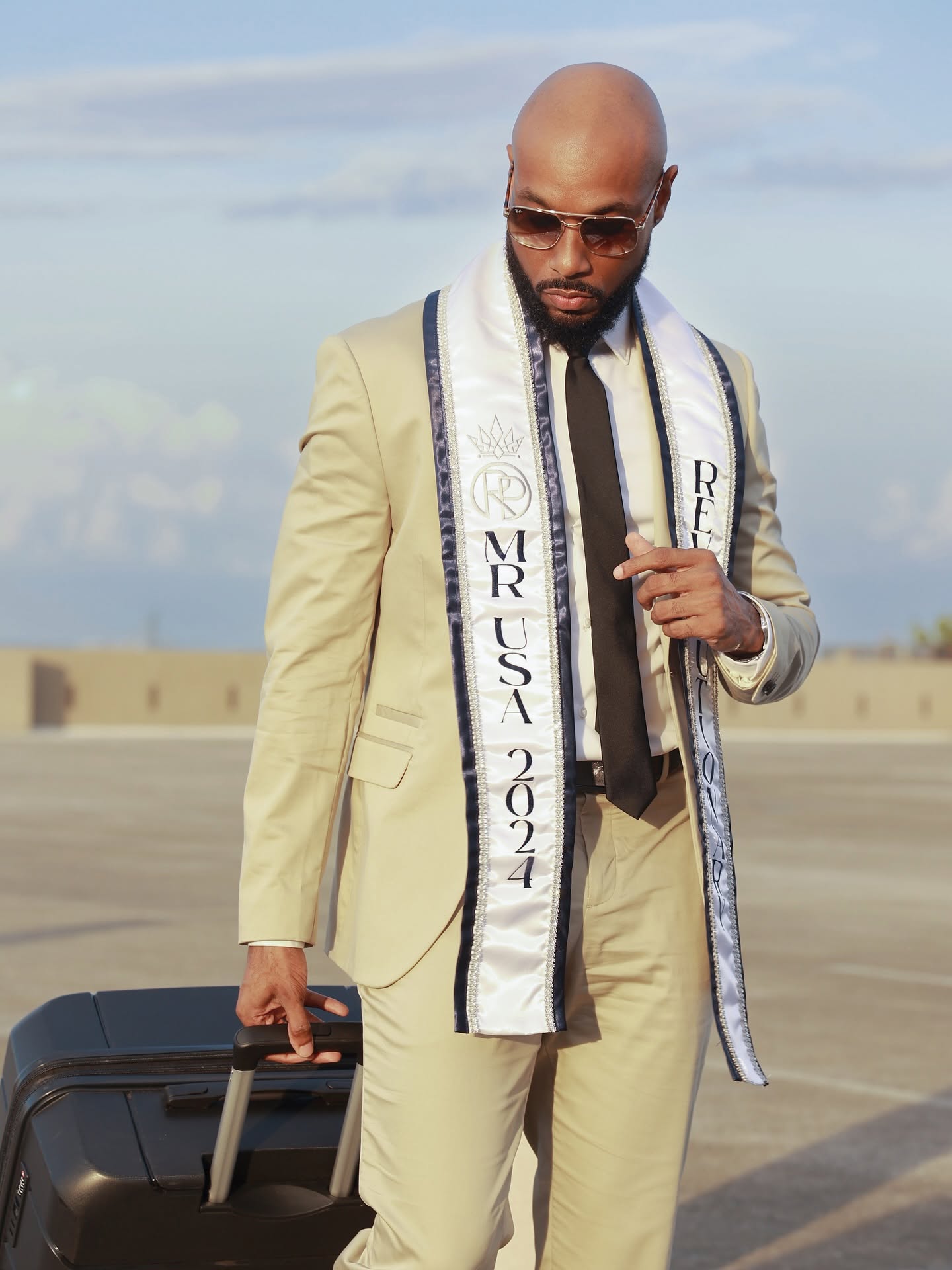 Man in beige suit with sunglasses holding a suitcase on an empty outdoor space, sky in background.