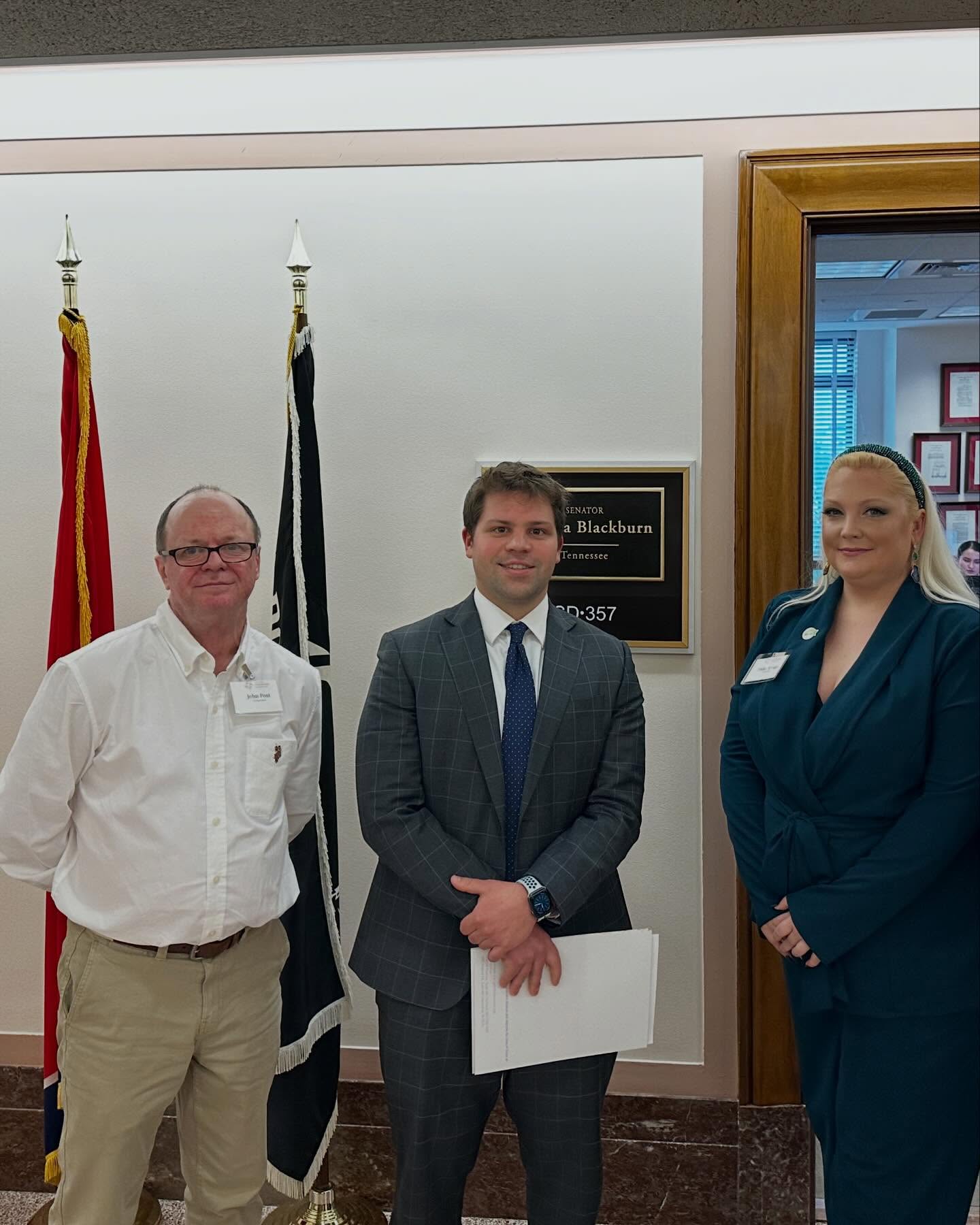 Three people standing indoors, two men and one woman, with flags and a plaque in the background.