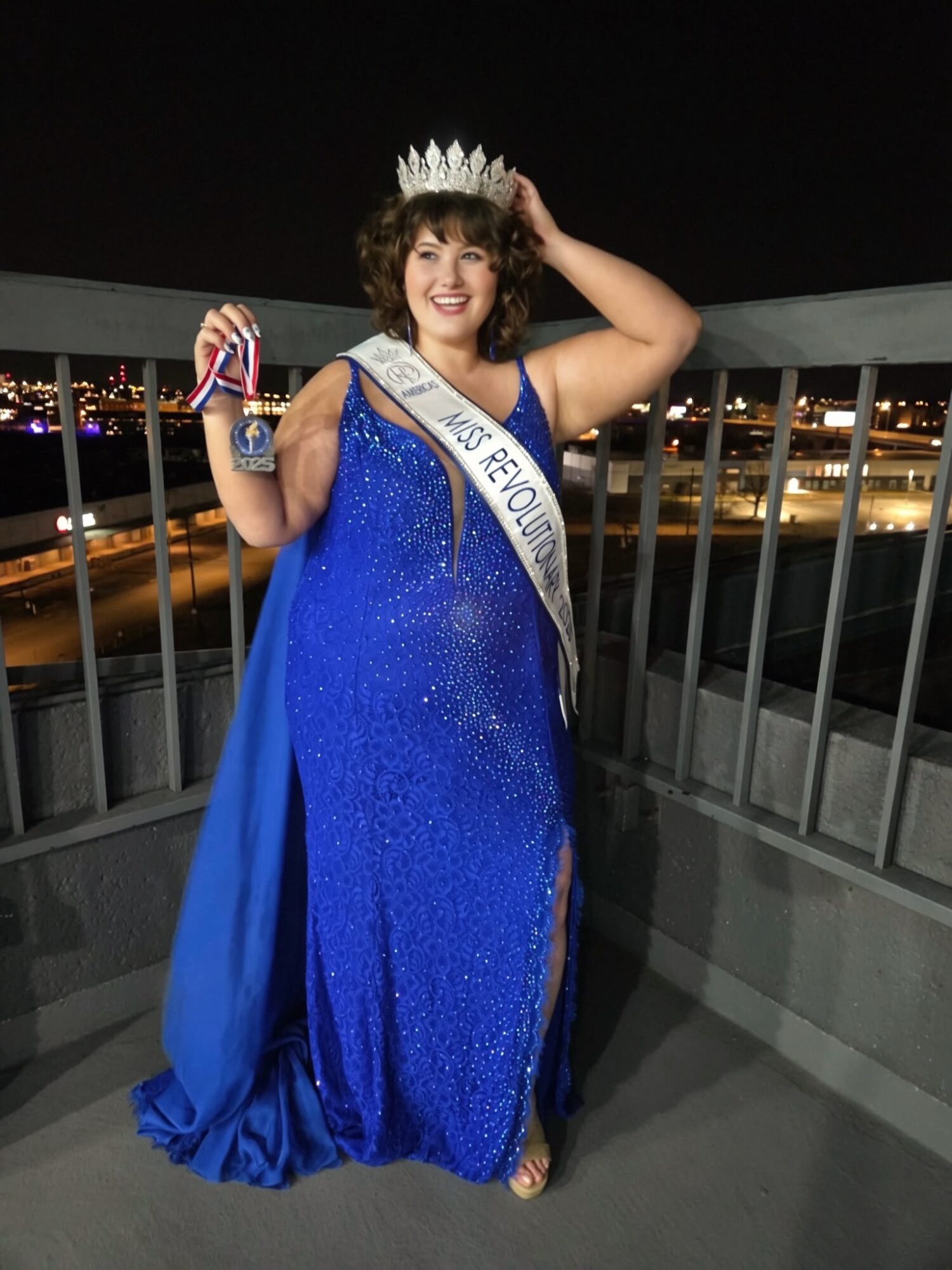 Woman in a blue gown and crown, holding a trophy, standing on a balcony at night with city lights in background.