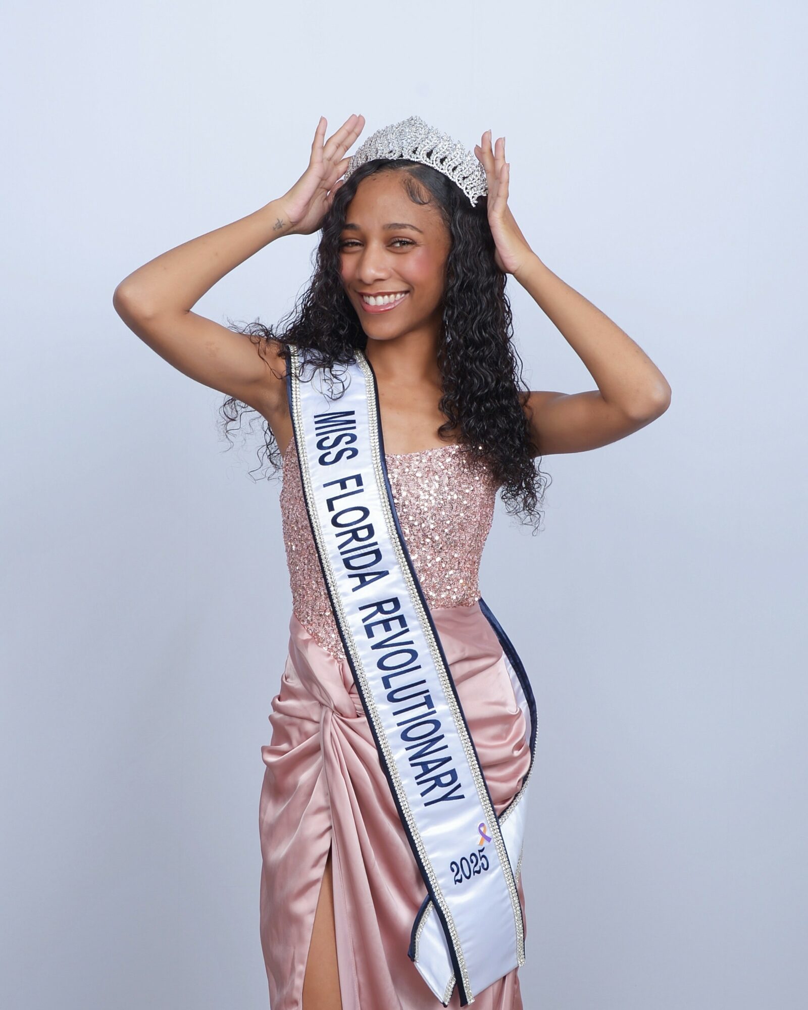 Young woman wearing a crown and sash, smiling, holding crown with both hands, against a plain light background.