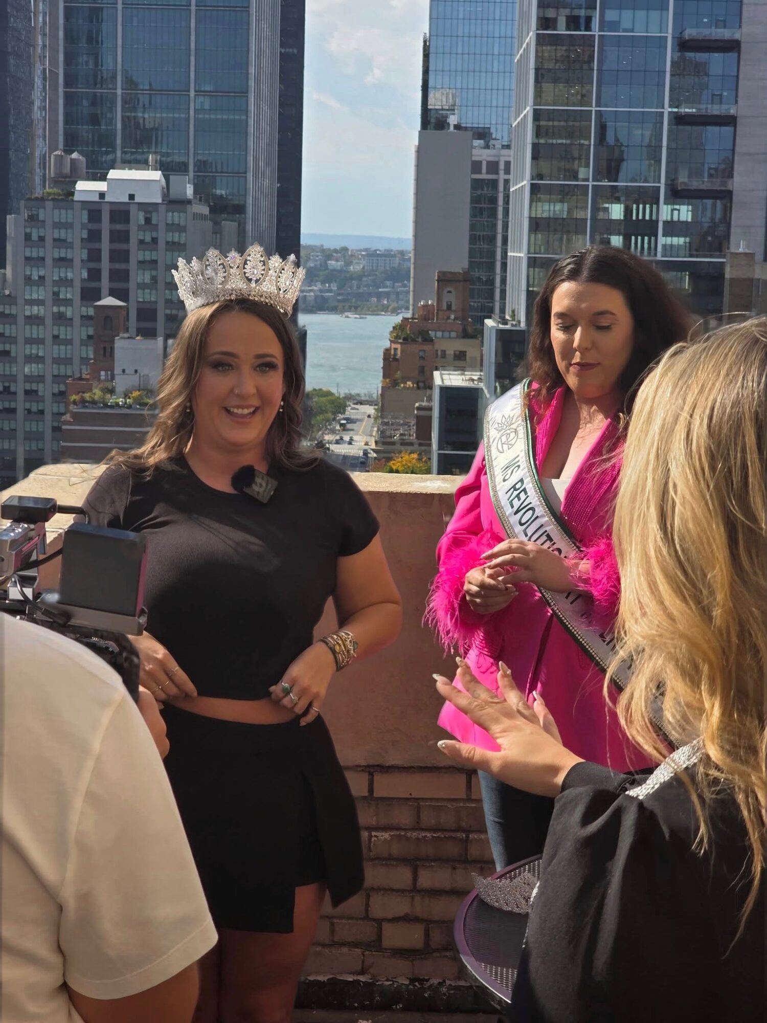Two women on a rooftop with city skyscrapers in the background, one wearing a crown and sash, smiling, the other in a pink outfit, holding a sash.