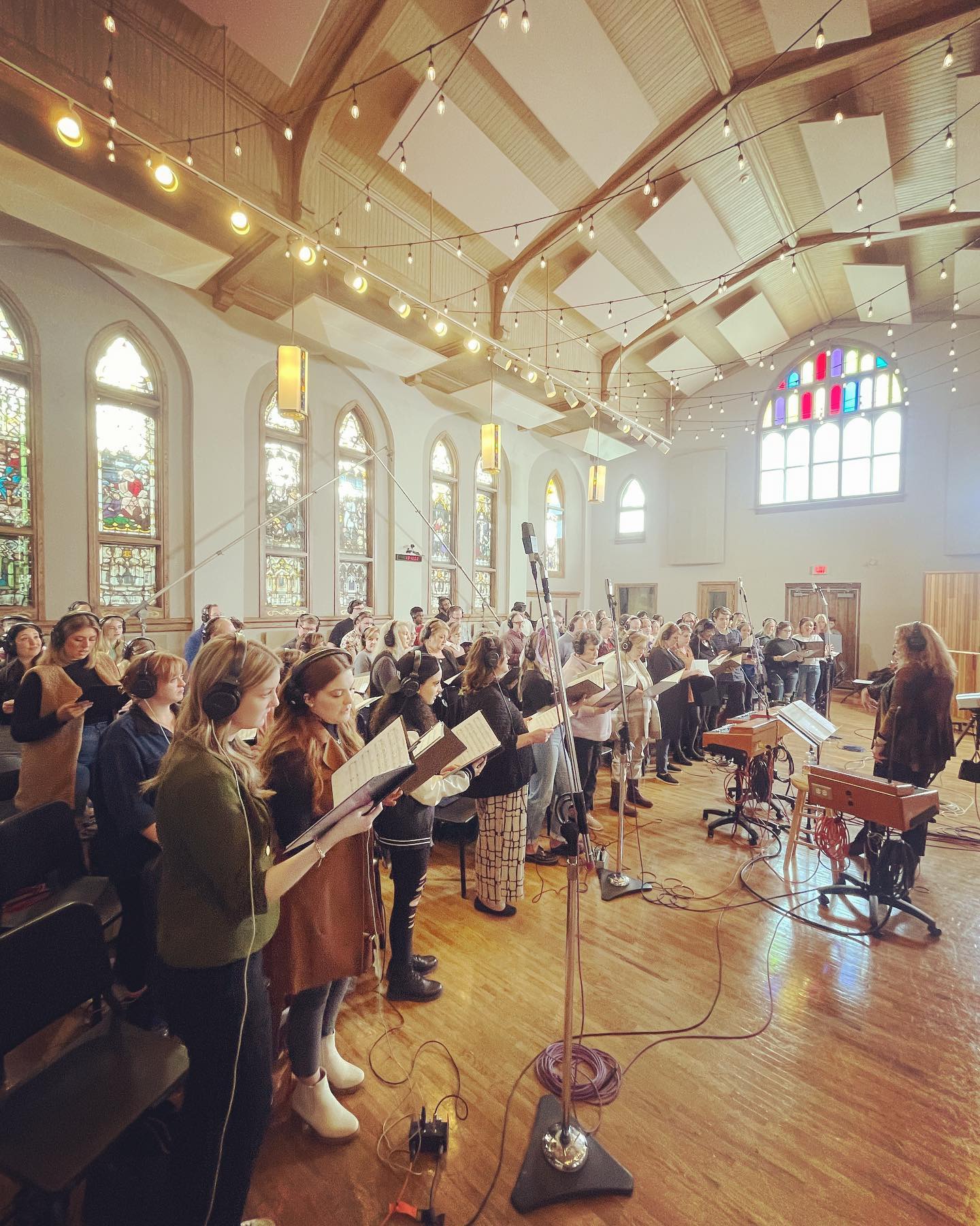 Group of people singing or rehearsing in a church or concert hall with large stained glass windows and a conductor.