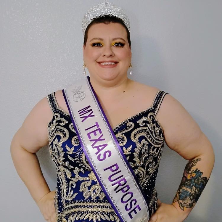 Woman wearing a crown and sash, smiling, in an ornate dress, with a tattooed arm, standing against a plain background.