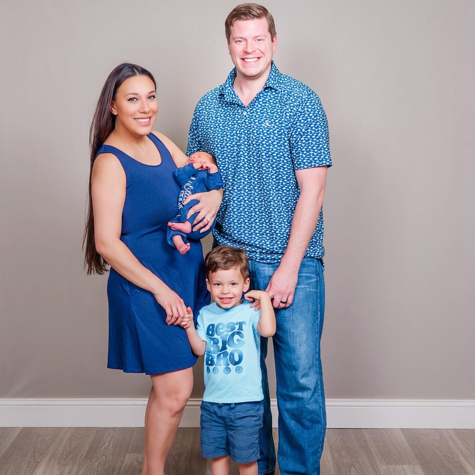 Family of three with a baby, standing indoors against a plain wall, smiling, with the woman holding the baby and the man standing beside them.