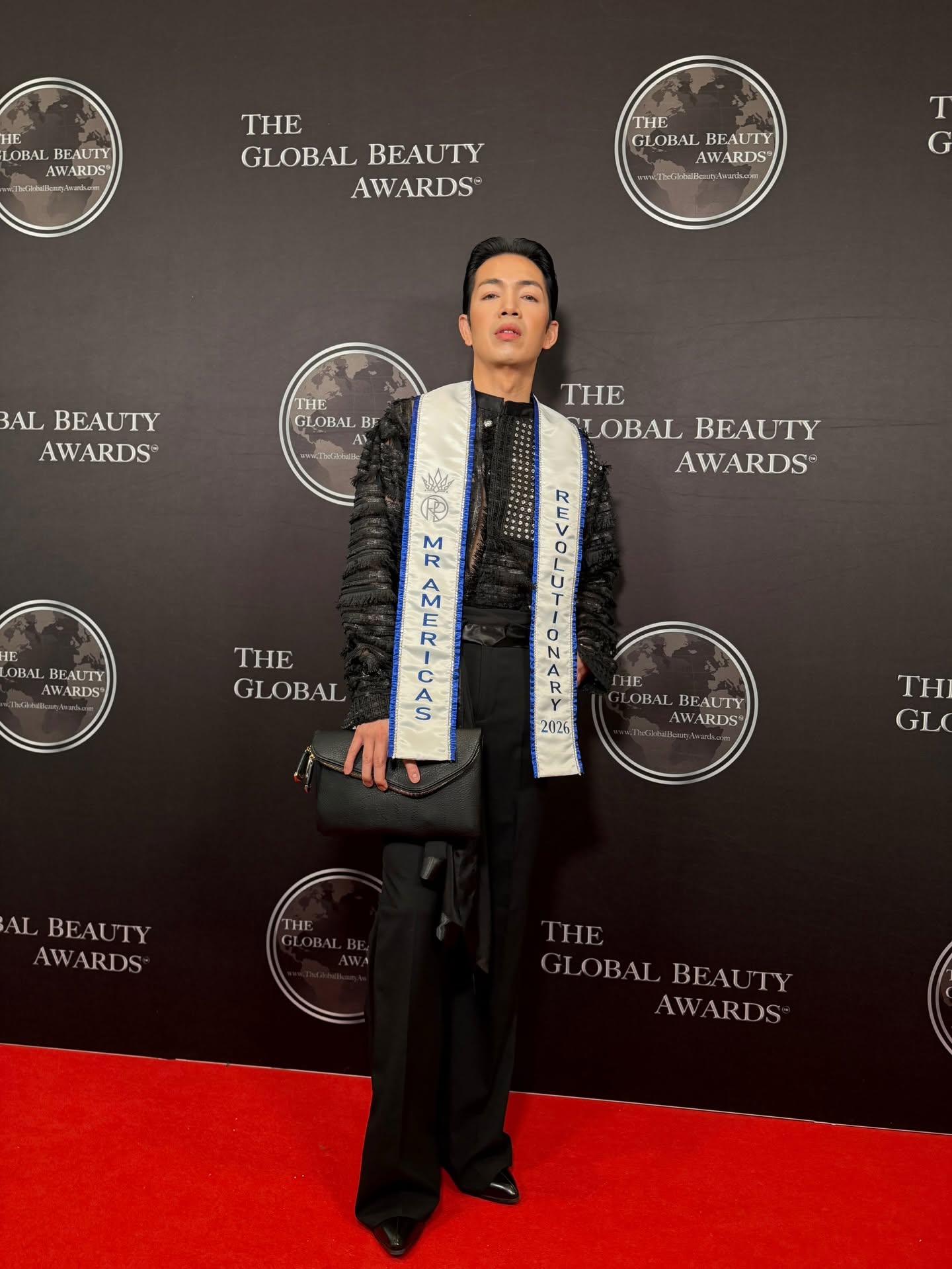 Person standing on red carpet in front of black backdrop with event logos, wearing a black outfit and sash, hands at sides.