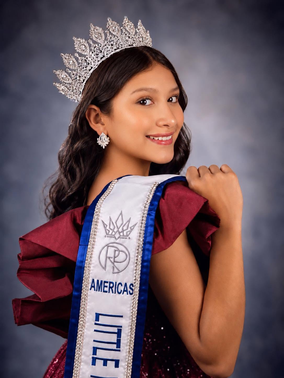 Young woman wearing a crown and sash, smiling, with dark wavy hair and earrings, against a dark background.
