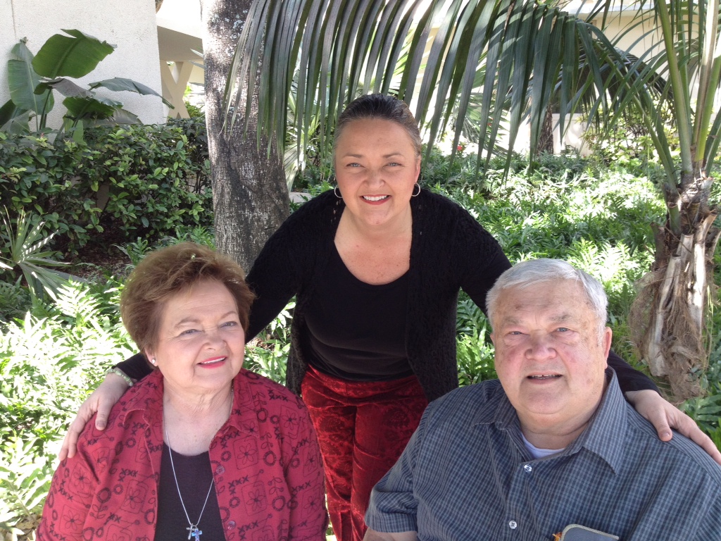 Three people smiling outdoors with greenery and palm trees in the background.