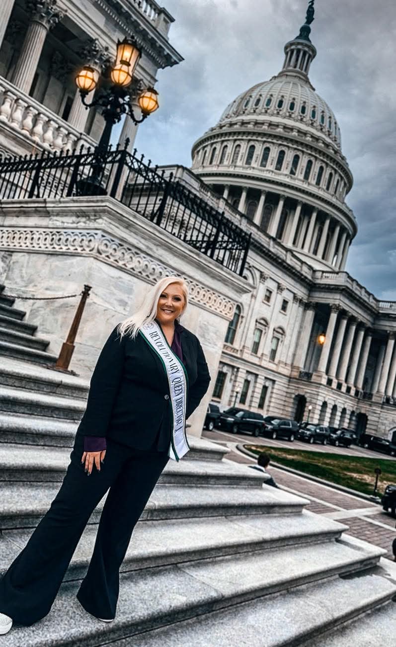 Woman in business attire standing on steps outside Capitol building with dome and cloudy sky in background.