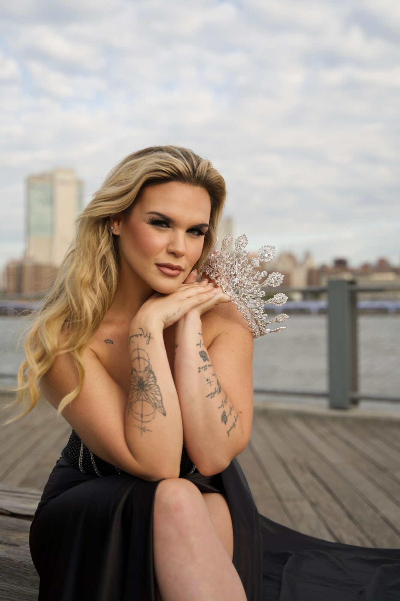 Woman with long blonde hair sitting outdoors near water, city skyline in background, wearing a black dress, resting her chin on her hands.
