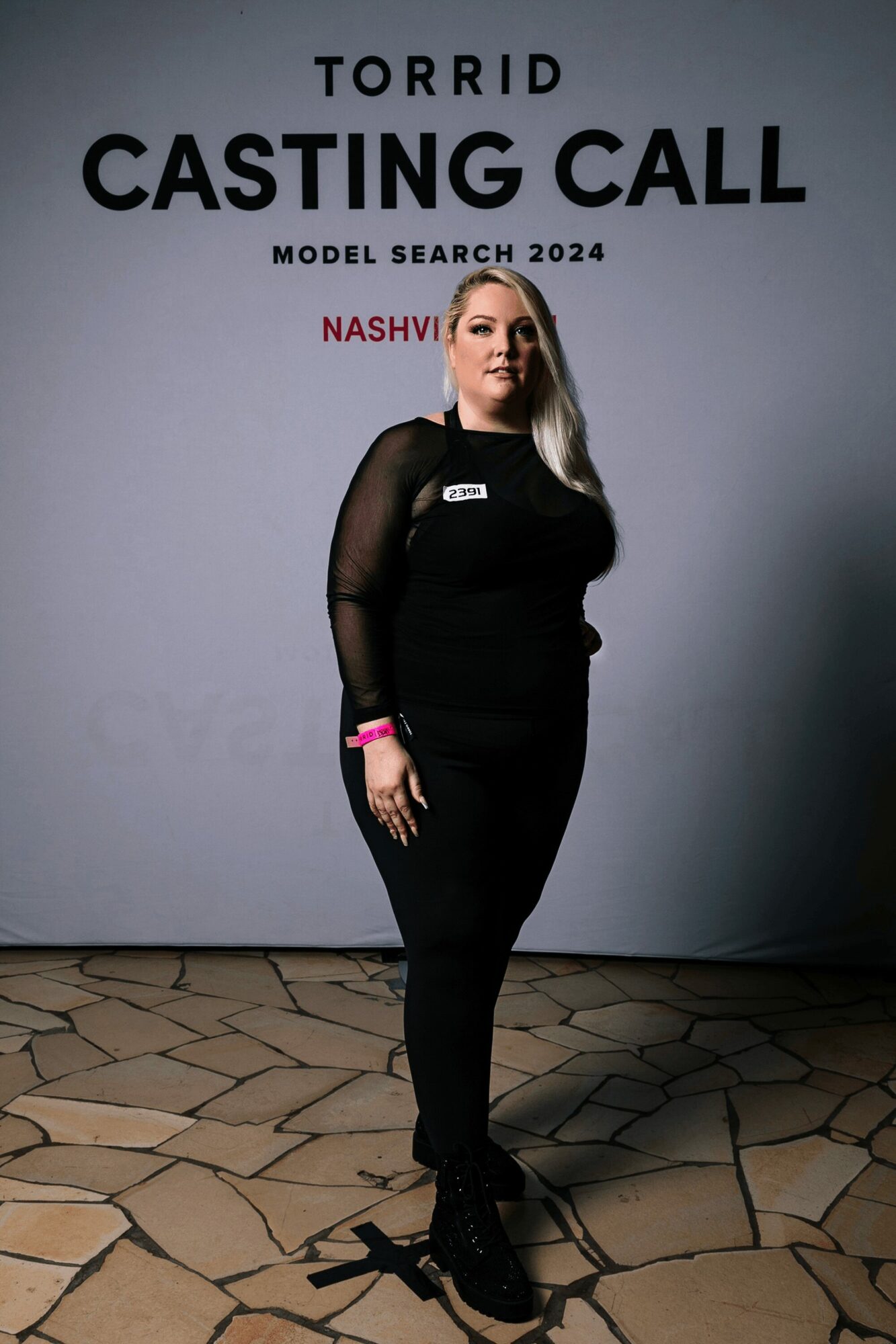 Woman standing in front of a casting call backdrop, wearing black clothing, with text behind her, on a patterned floor.