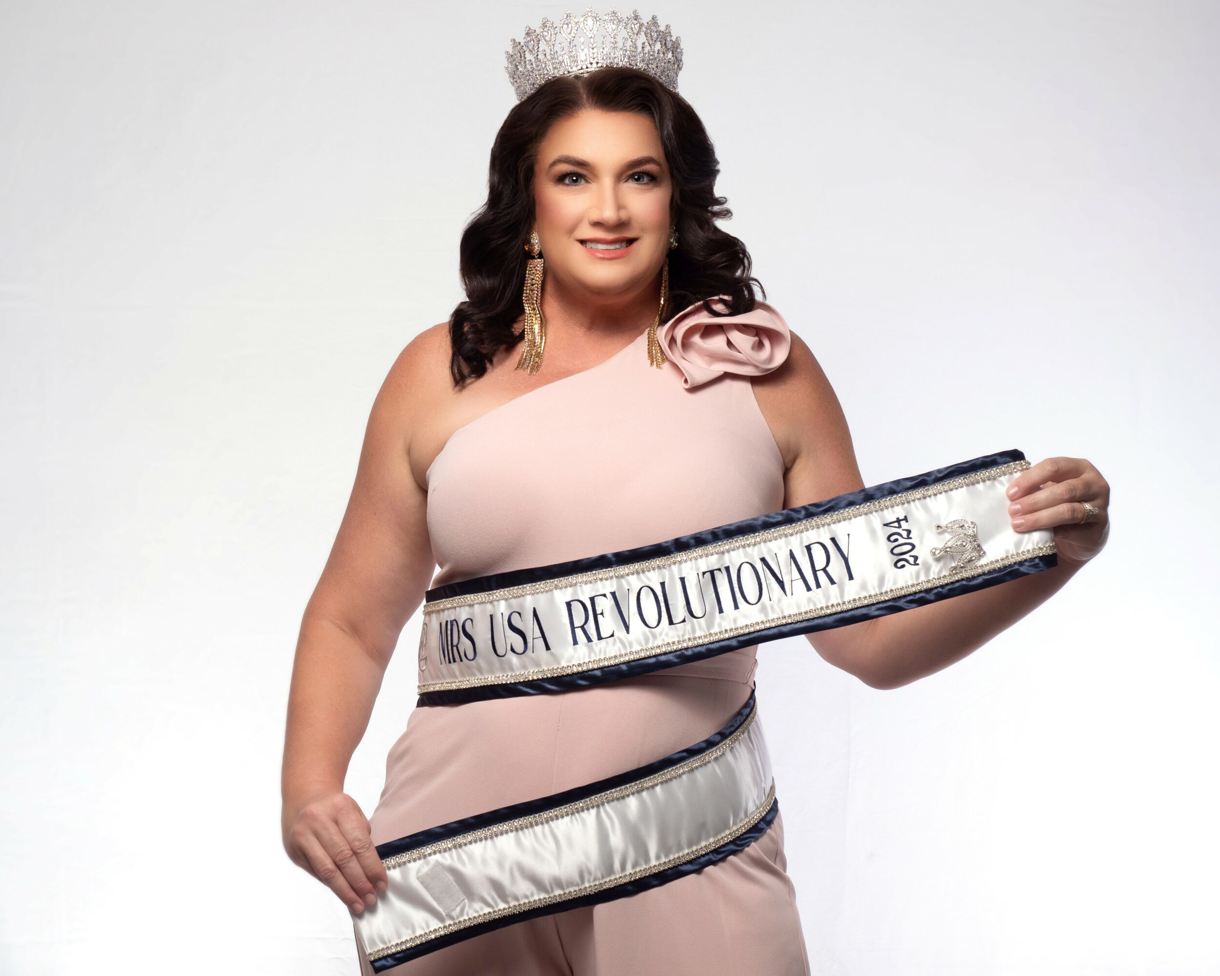 Woman wearing a crown and sash, holding a large circular object, smiling, against a plain background.
