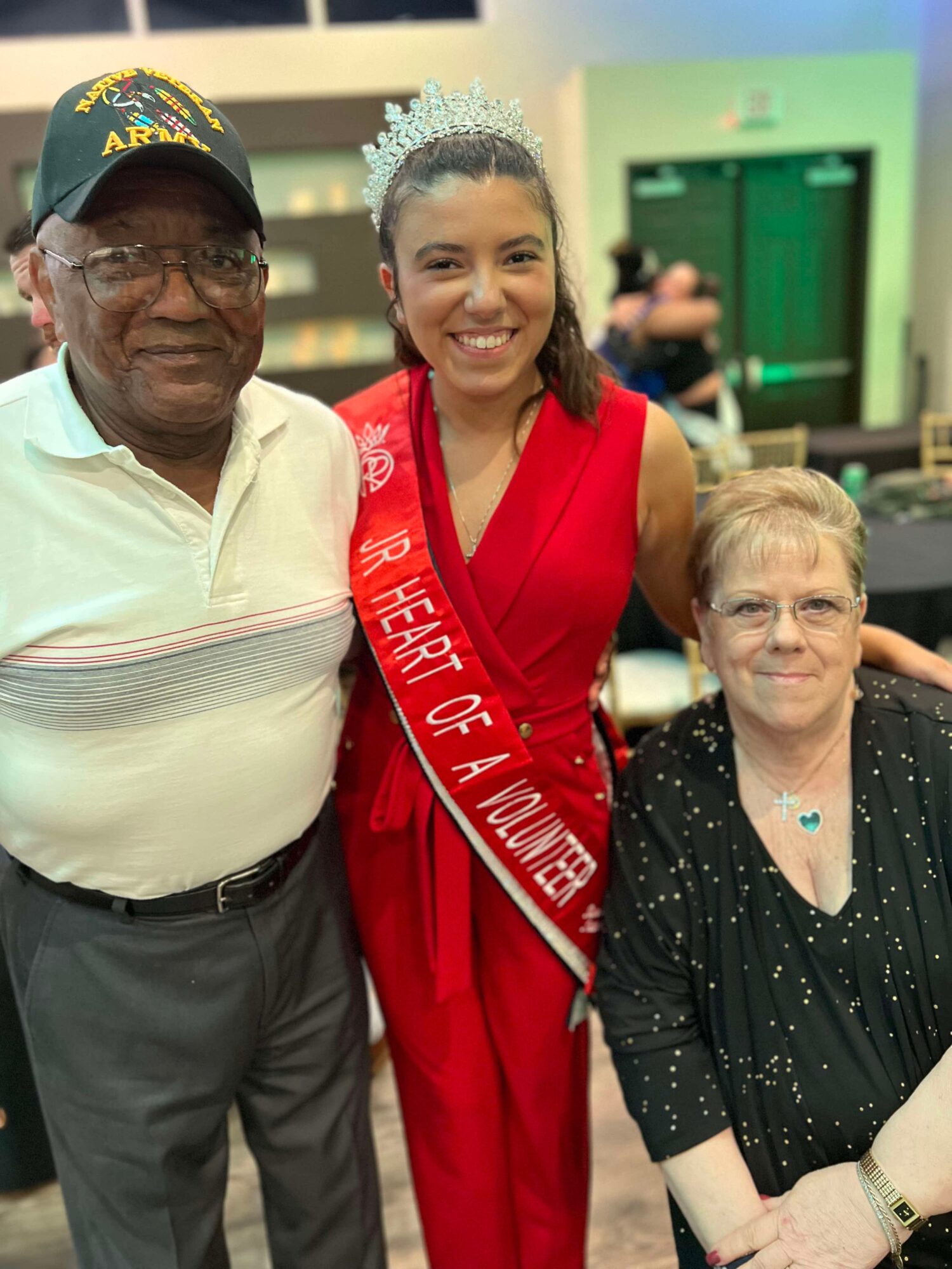 Young woman wearing a crown and sash, smiling, with two older adults, one man and one woman, in an indoor setting.