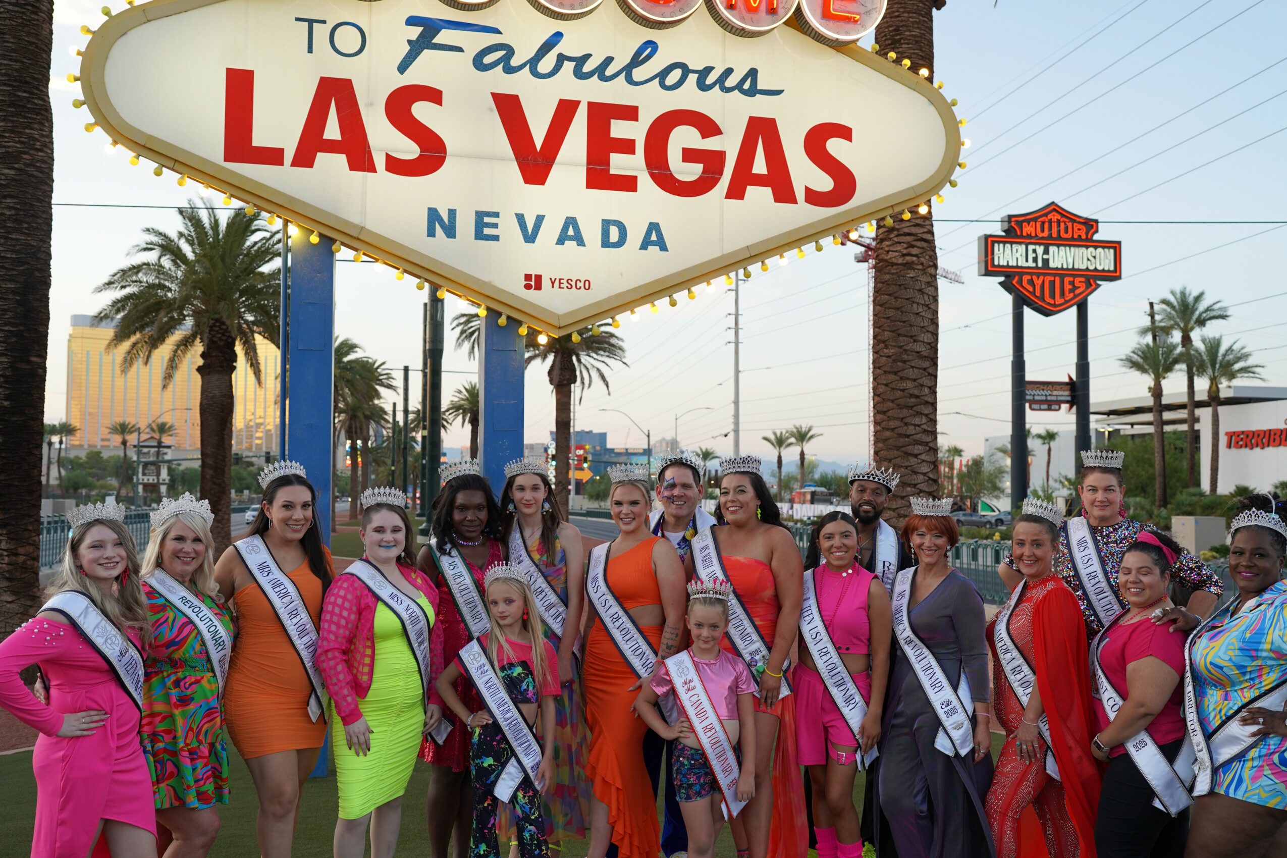 Group of women in colorful dresses and sashes standing in front of Las Vegas sign with palm trees and buildings in background.