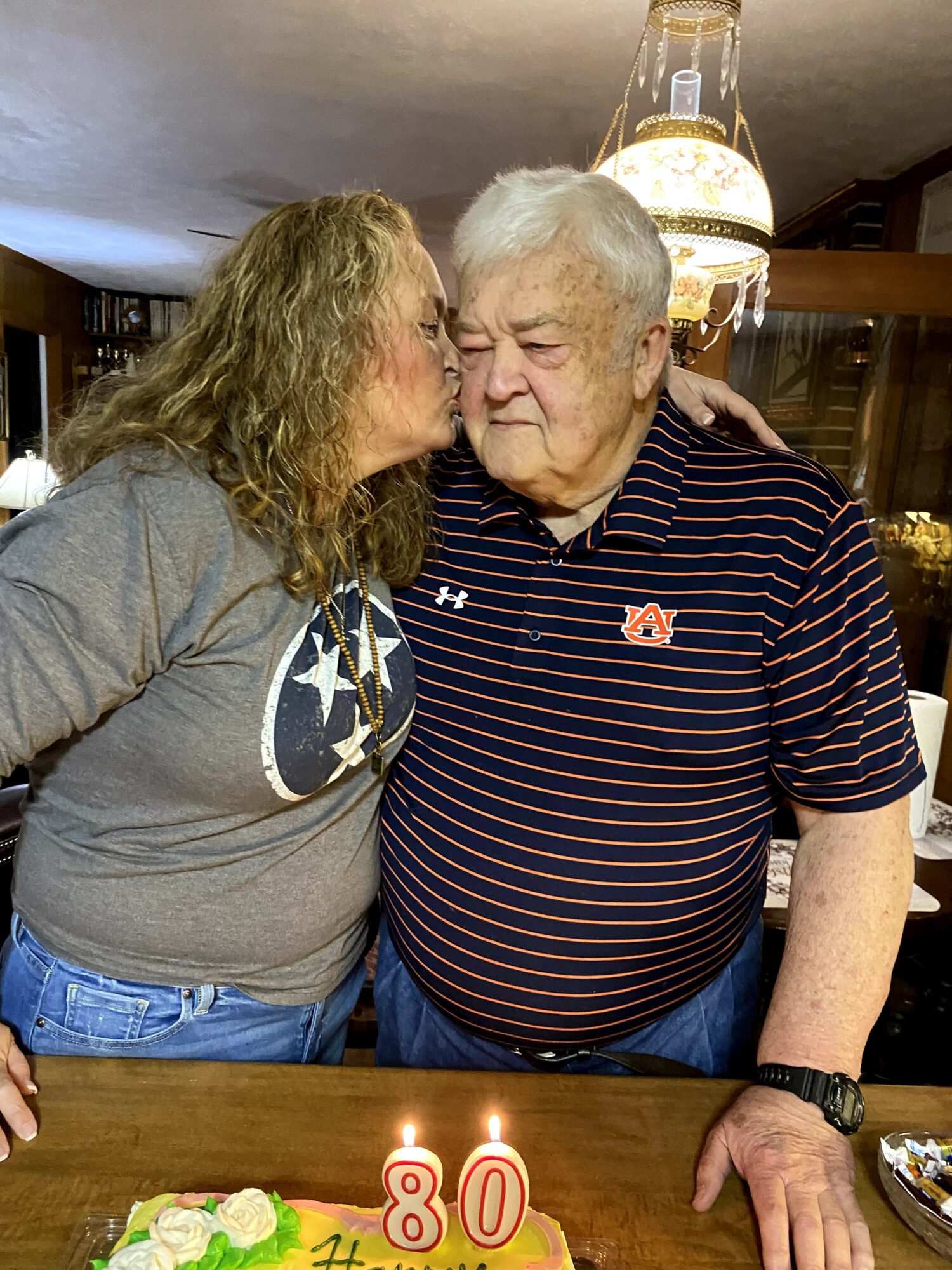 Woman kisses elderly man on the cheek in a room with a lit birthday cake and candles, celebrating an 80th birthday.
