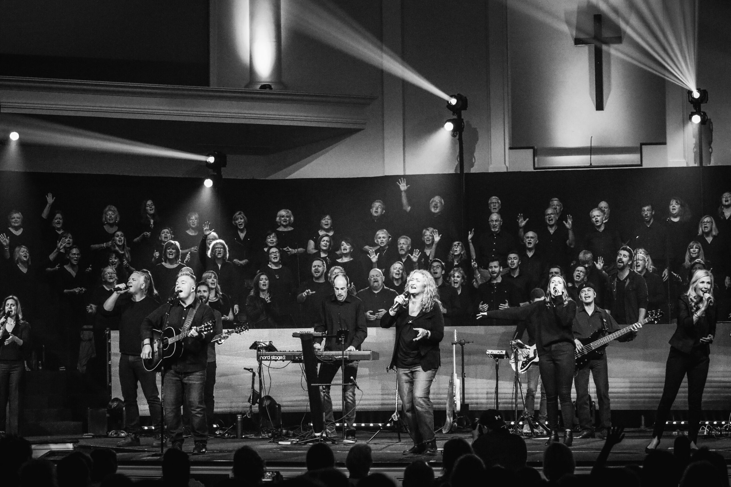 Group of musicians and singers performing on stage with lighting and a cross in the background.