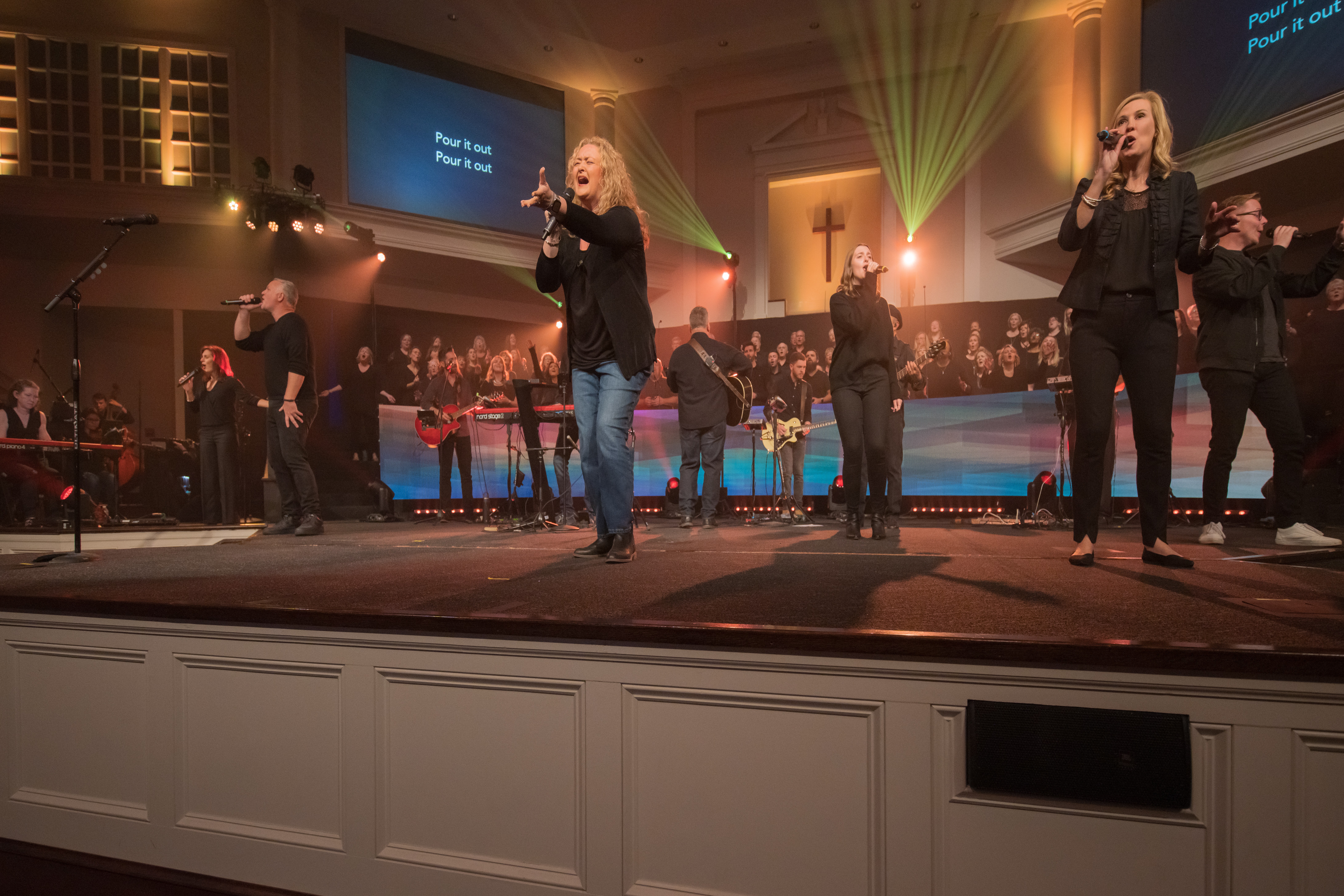 Group of people singing and playing instruments on stage in a church setting with a choir in the background.
