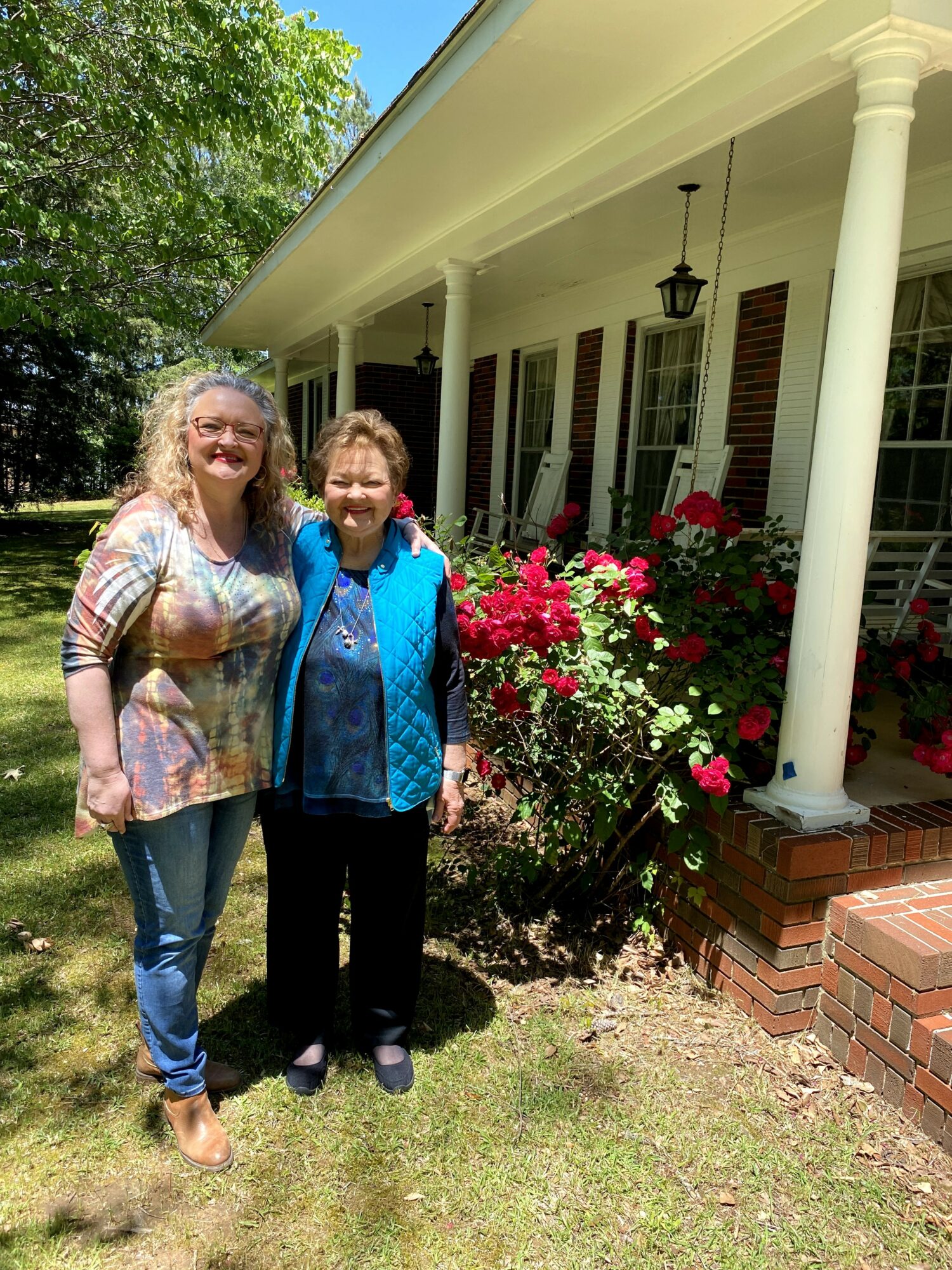 Two women stand outside a house with white columns and pink flowers, smiling at the camera.
