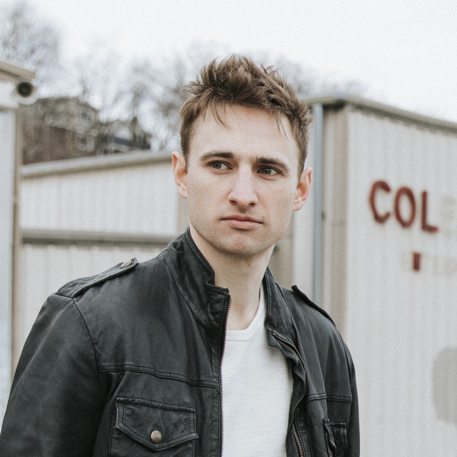 Young man with short brown hair wearing a black jacket and white shirt, outdoors near storage containers.
