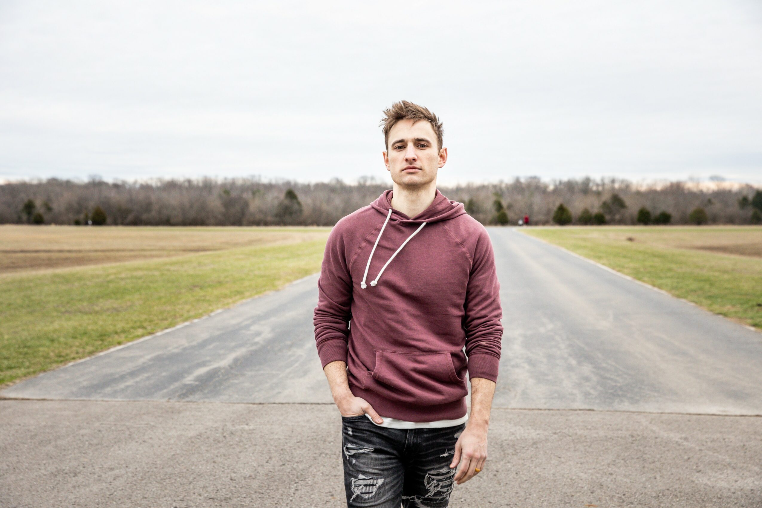Young man standing on a road in an open field, wearing a maroon hoodie and jeans, with a neutral expression.