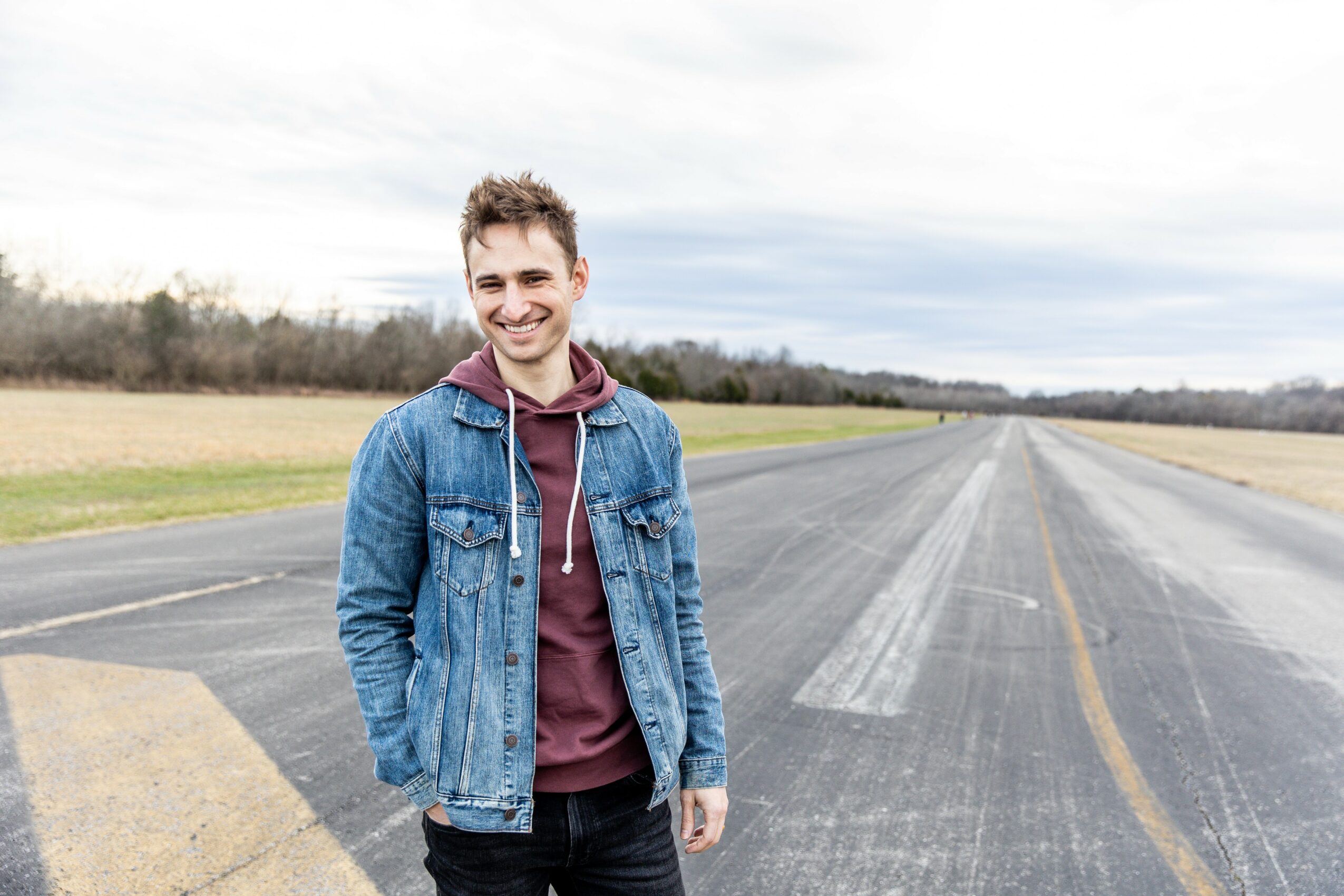 Young man smiling, standing on an empty road with open fields and cloudy sky in background.