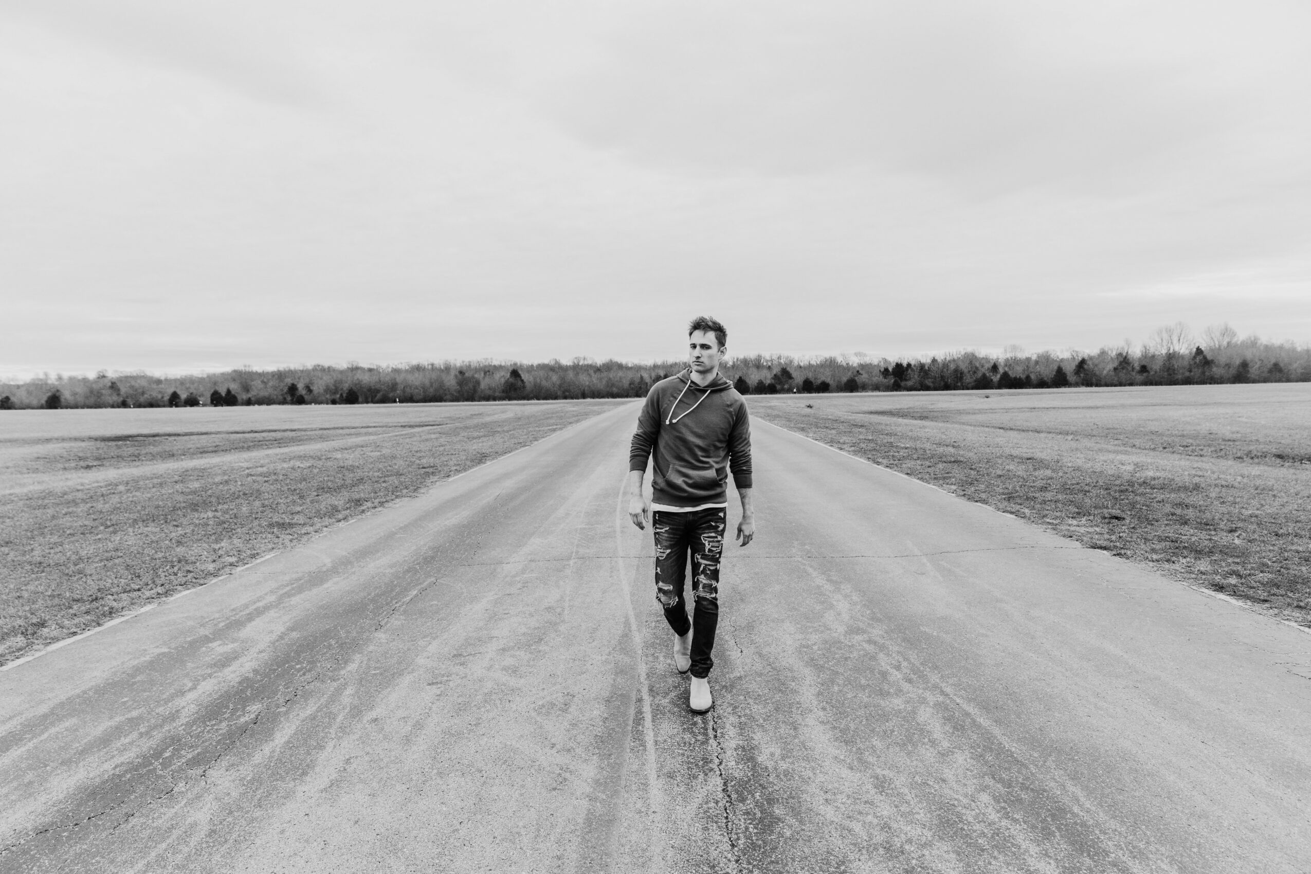 Person walking on a straight road in an open field under a cloudy sky, holding a leash, in black and white.