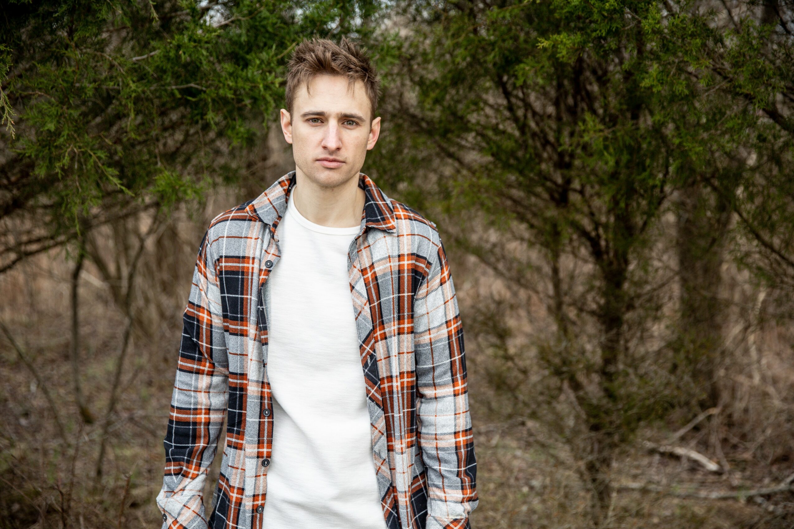 Young man standing outdoors with trees in the background, wearing a plaid shirt over a white t-shirt.