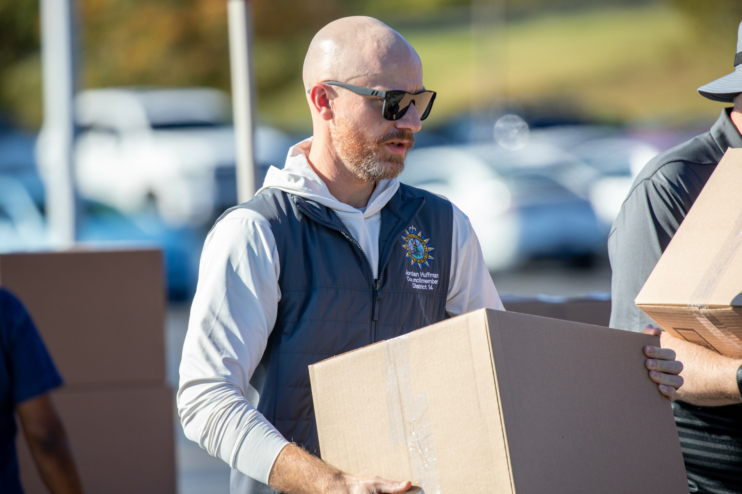 Man with sunglasses and a beard holding a cardboard box outdoors, with cars and trees in the background.