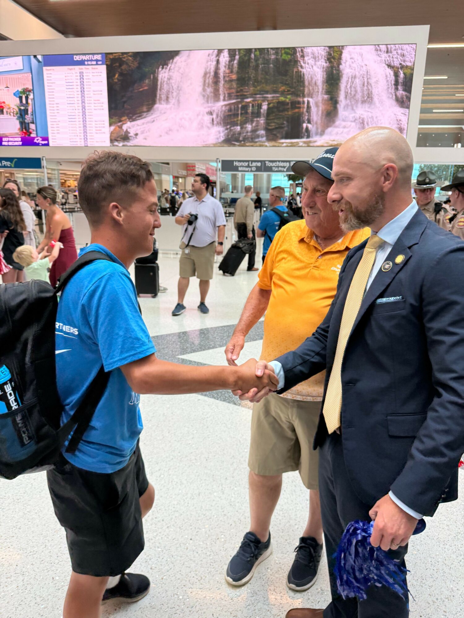 Two men shaking hands in an indoor public space with people and display boards in the background.