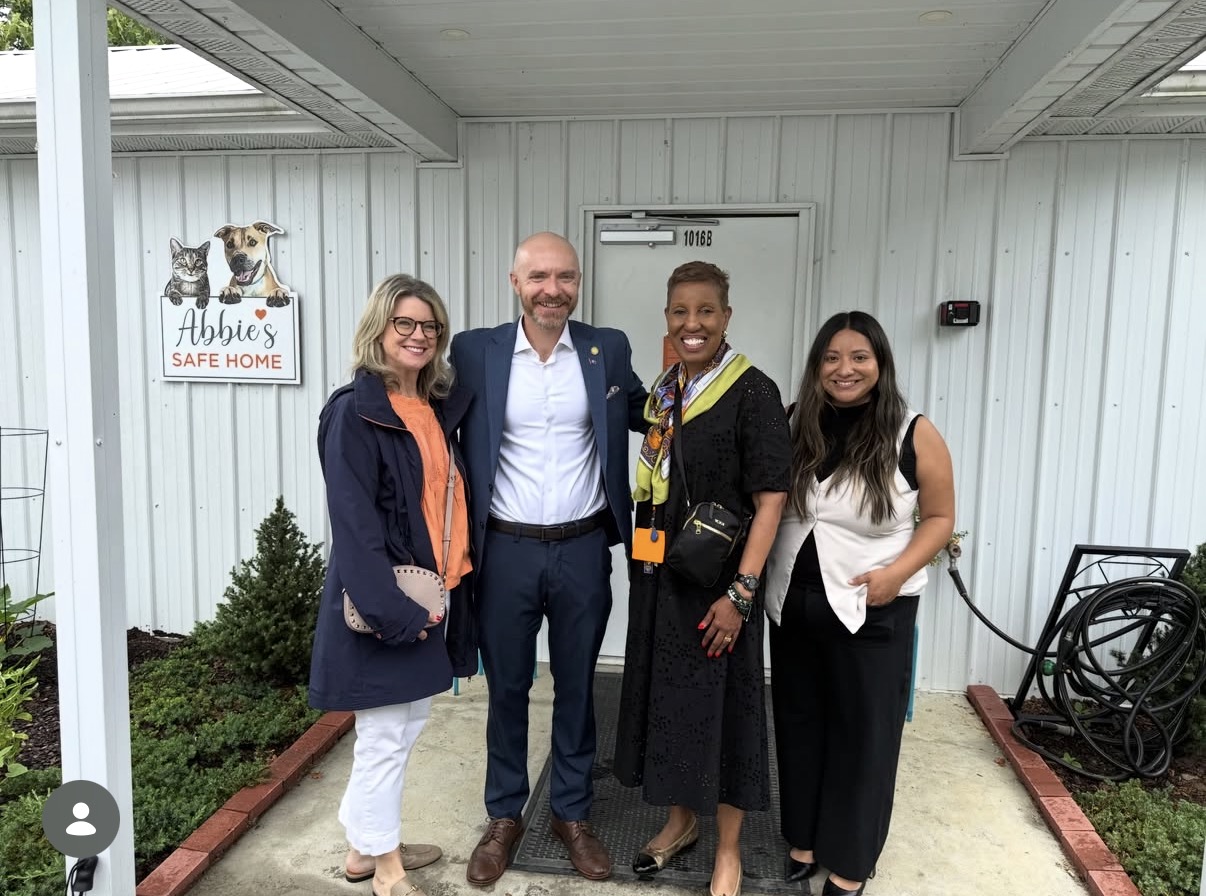 Four people standing outside a building, smiling, with a sign that reads 'Abbie's Safe Home' and a dog image, in front of a white wall.