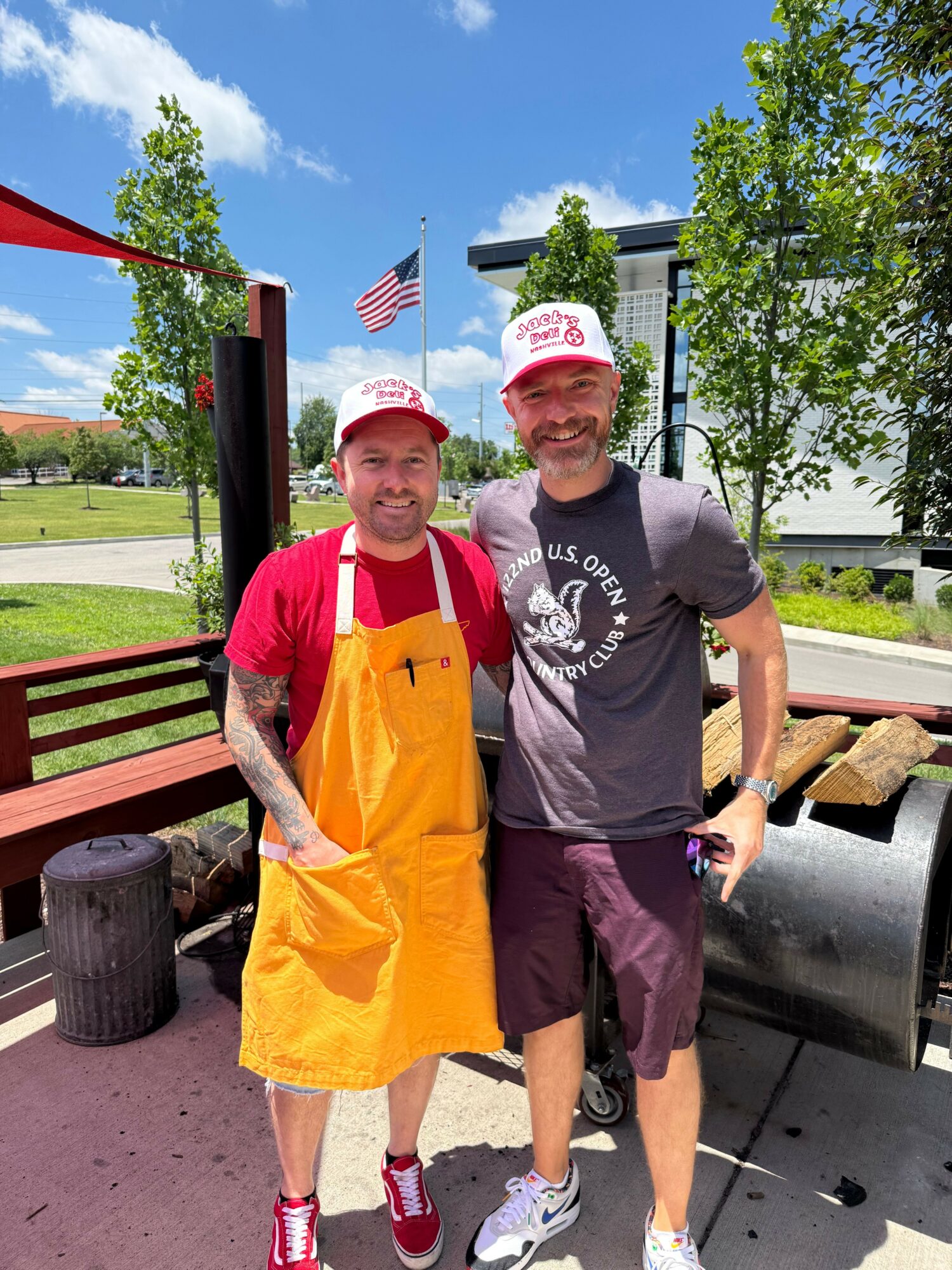 Two people standing outdoors, one in a gray shirt and shorts, the other in a red shirt and yellow apron, with trees and a blue sky in the background.