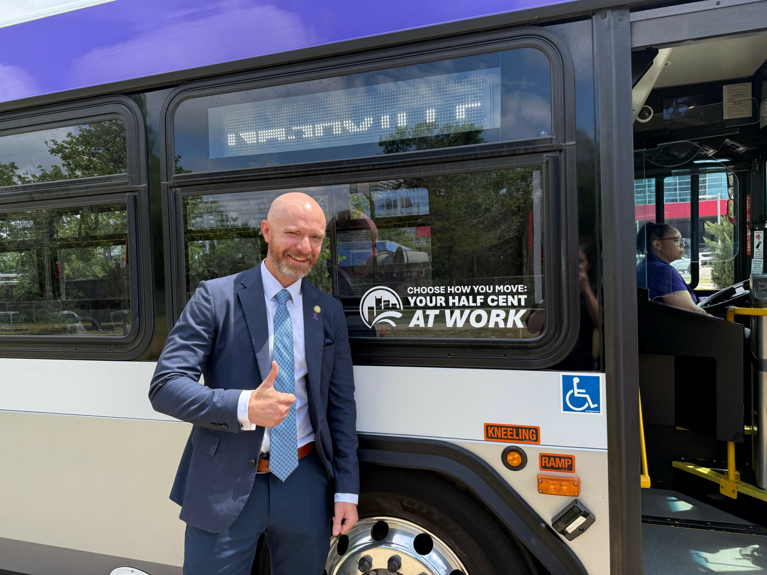 Man in suit giving a thumbs-up next to a bus with accessible signage, window reflections visible.