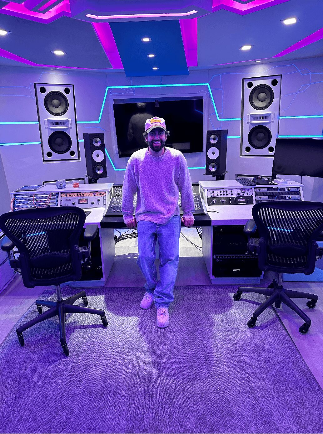 Person standing in a music studio with large speakers and audio equipment, illuminated by purple and blue lighting.