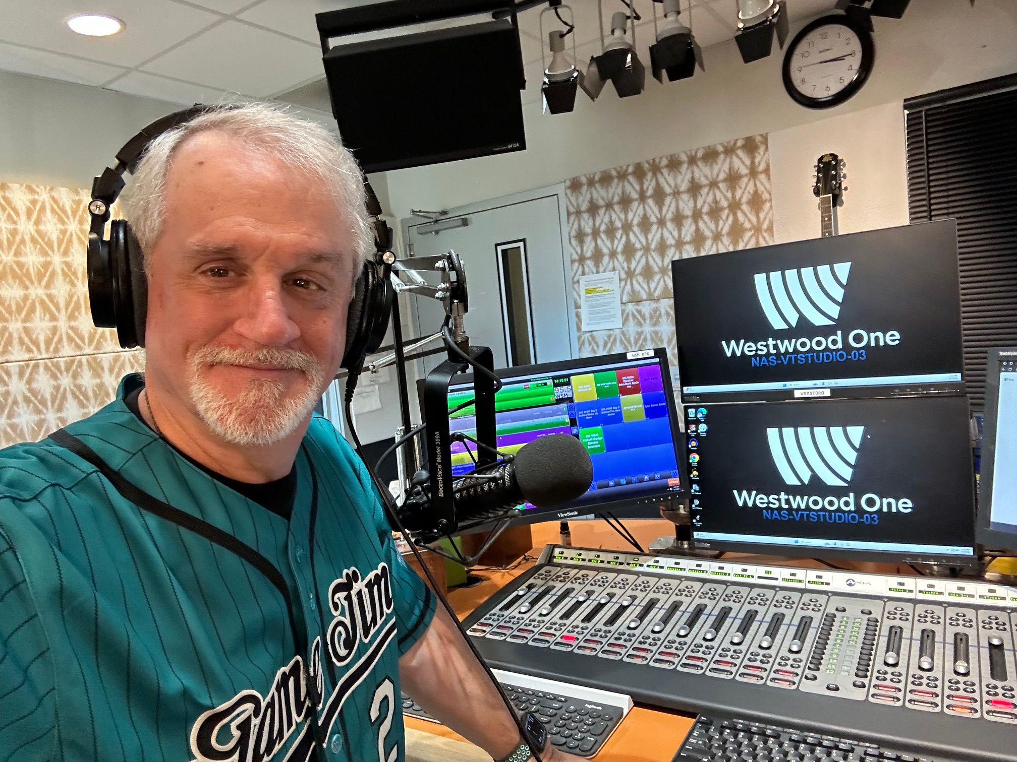 Person with headphones in a radio studio, microphone, sound equipment, and computer screens visible.