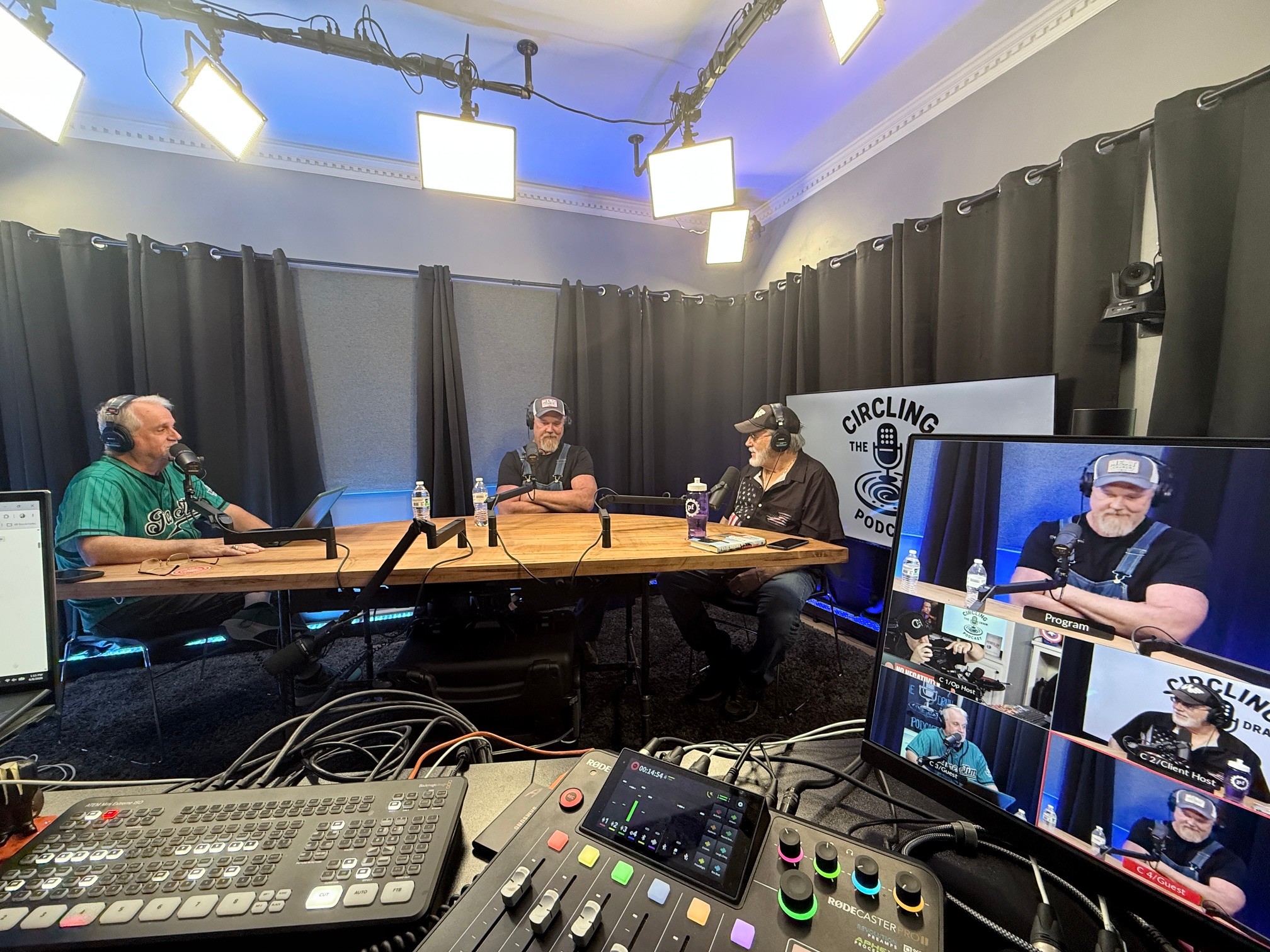 Four people sit around a table in a recording studio with black curtains, microphones, and a monitor showing the scene.