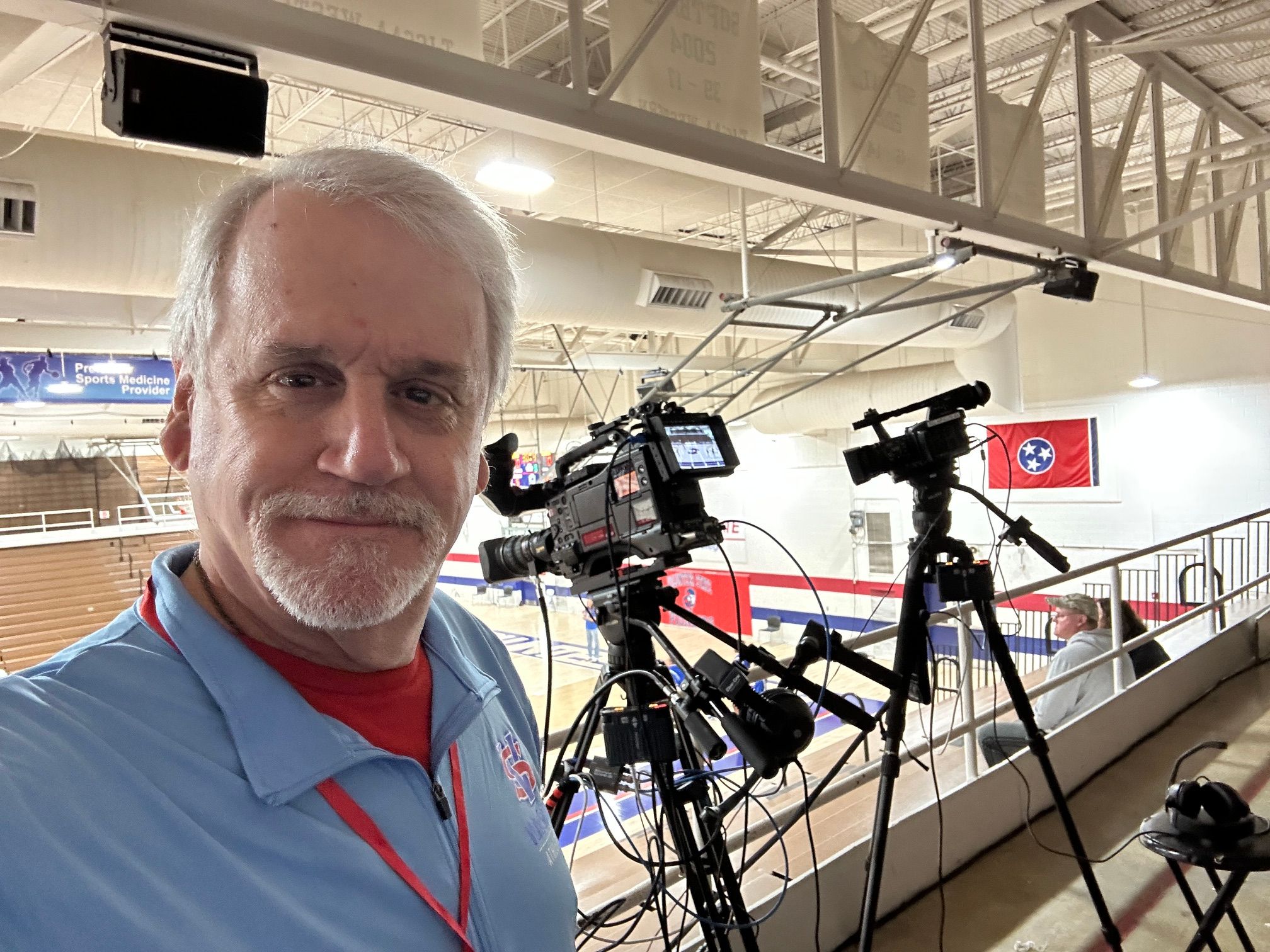 A man with gray hair and a beard takes a selfie in an indoor sports facility with a bicycle overhead.