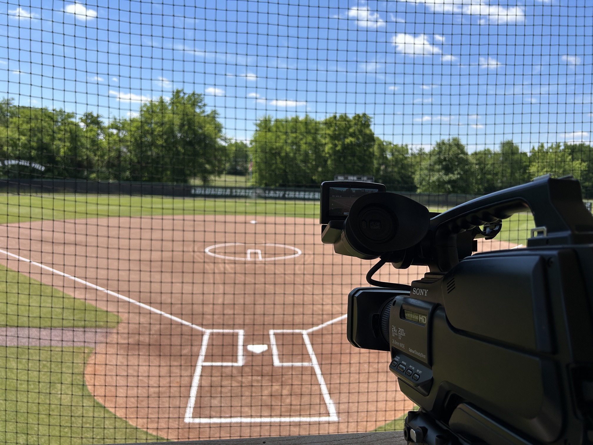 View of a baseball field with a camera in the foreground, netting, trees, and a blue sky with clouds.