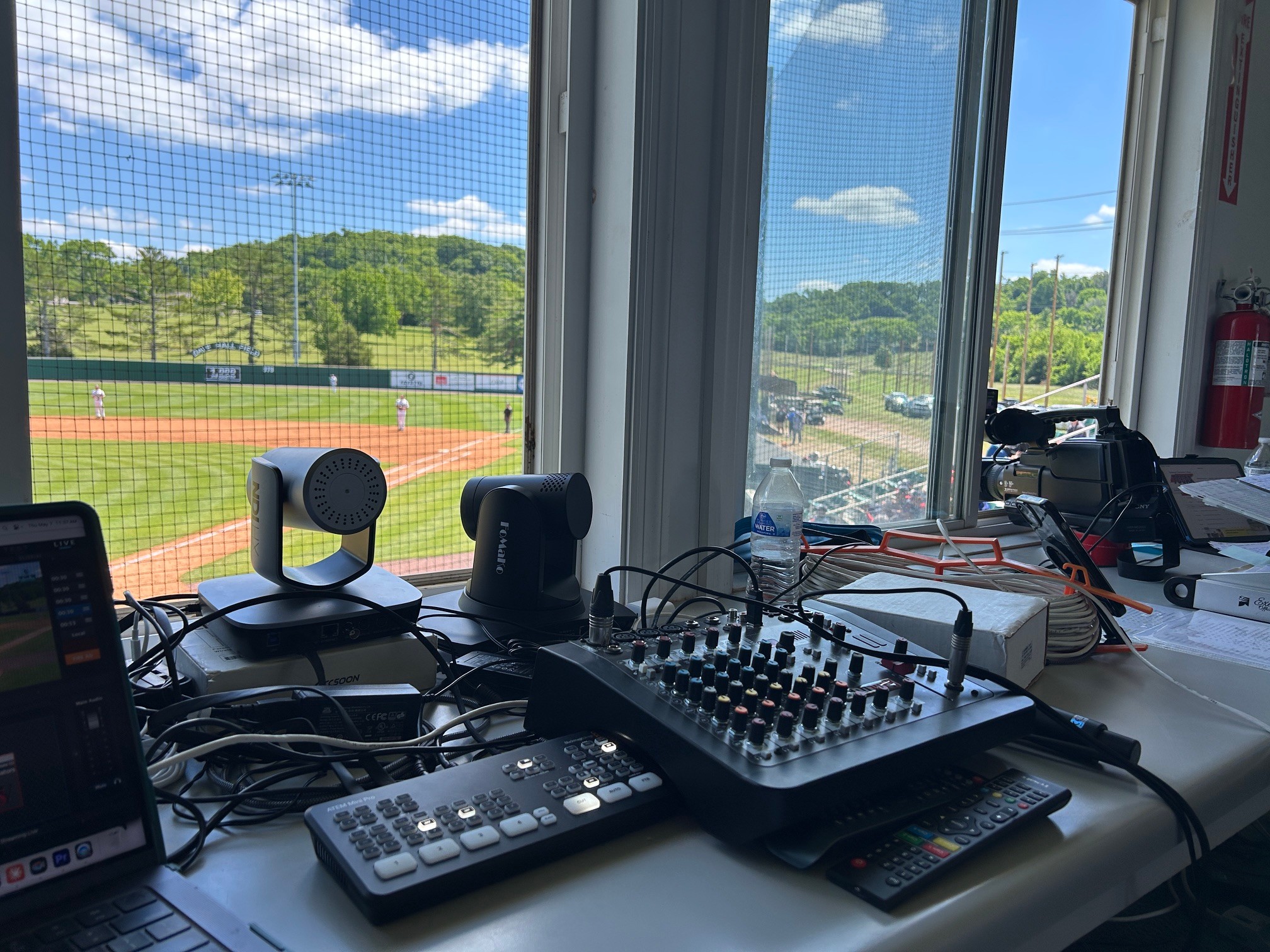 View from inside a room showing a table with audio equipment, a window with a view of a baseball field and green landscape, blue sky, and clouds.