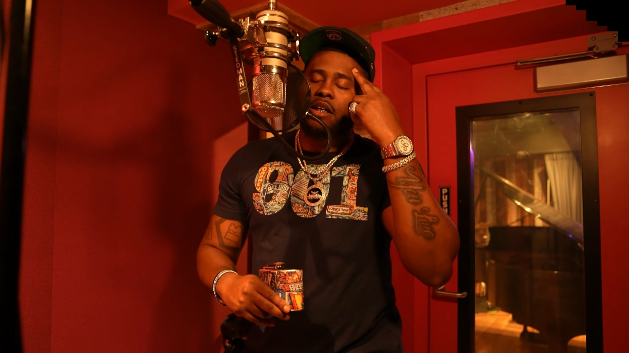 Man in a recording studio holding a glass, wearing a black t-shirt with colorful text, and jewelry.
