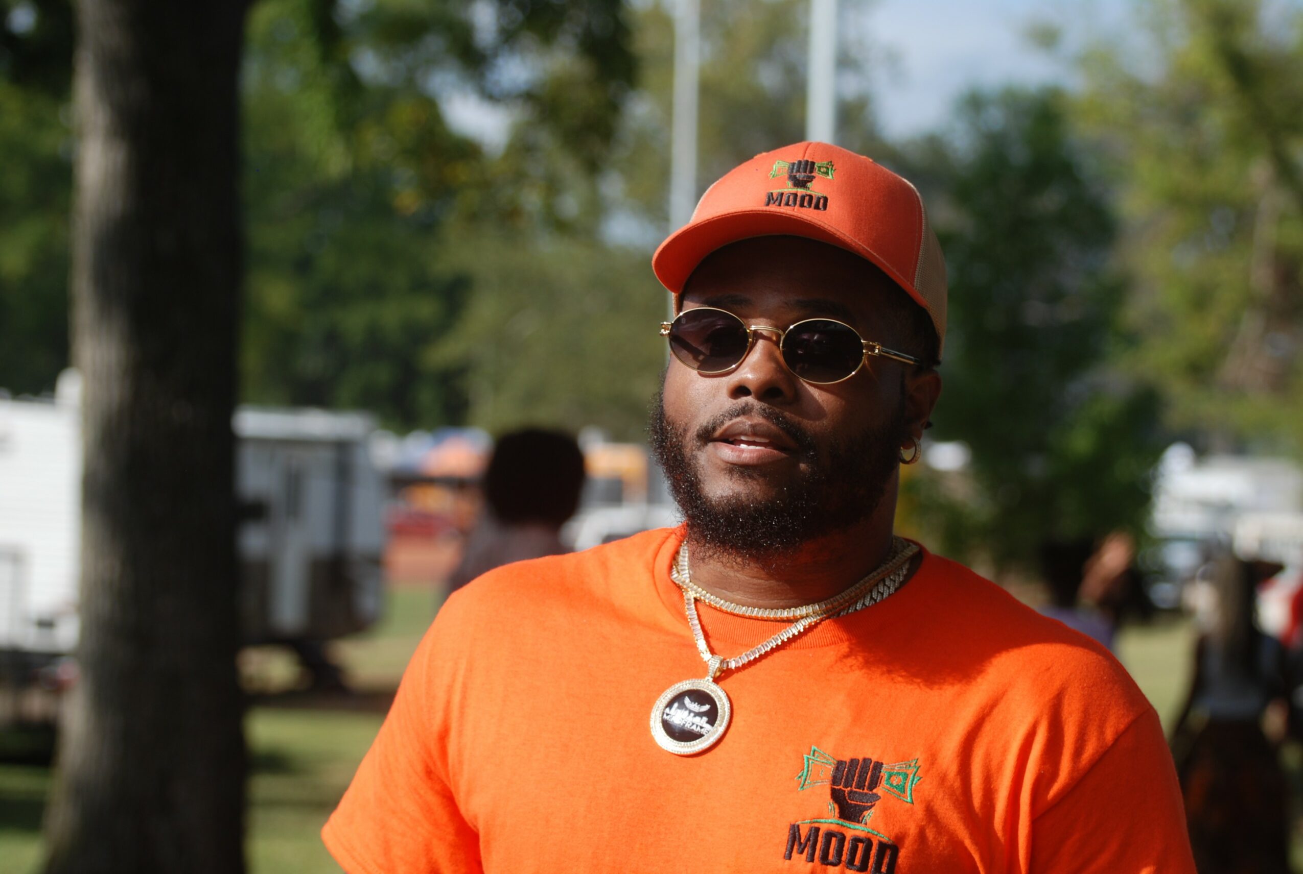 Man wearing sunglasses, orange cap, and orange shirt outdoors with trees and people in background.