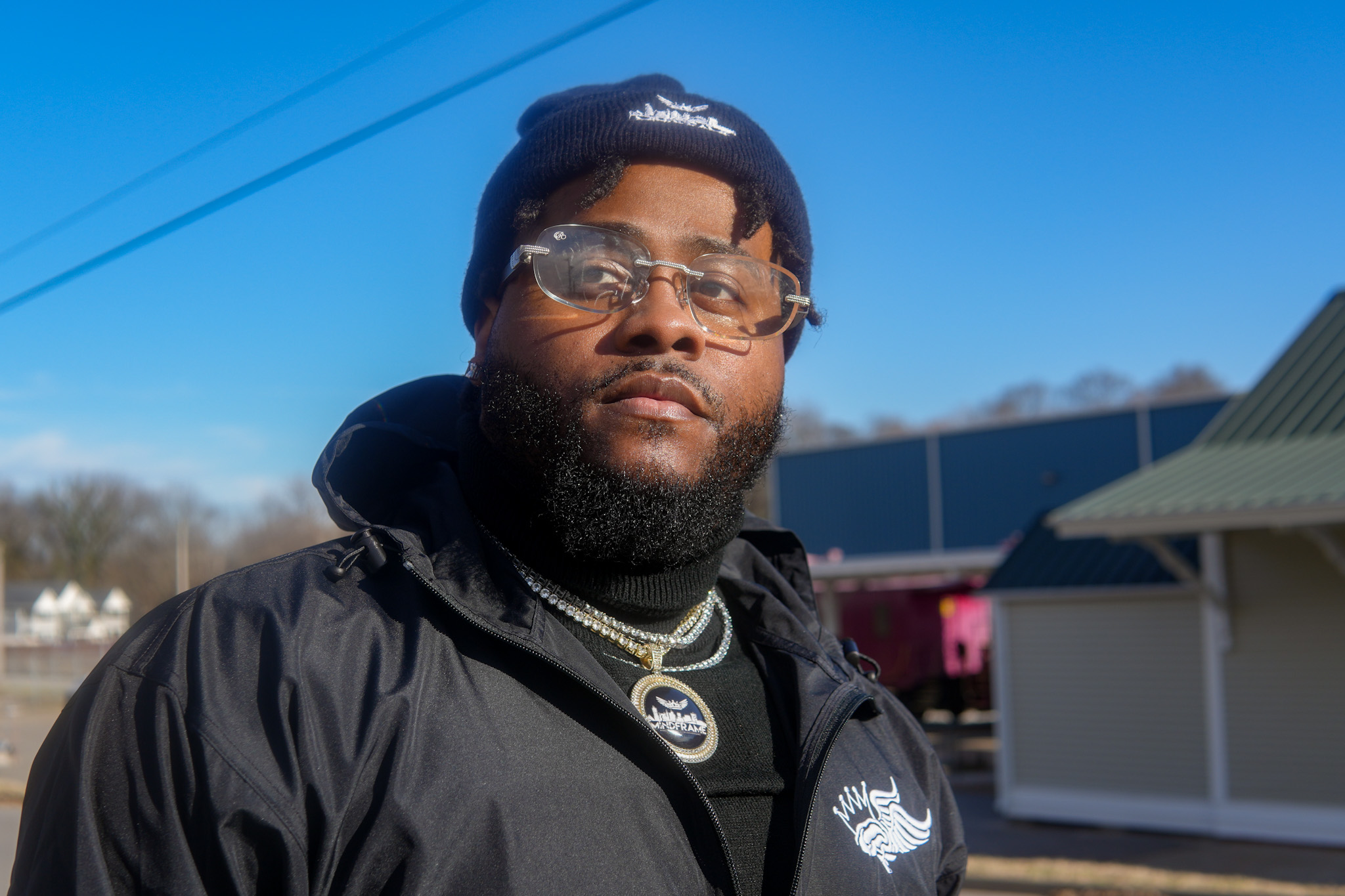 Man with glasses and a beanie, wearing a black jacket outdoors on a sunny day.