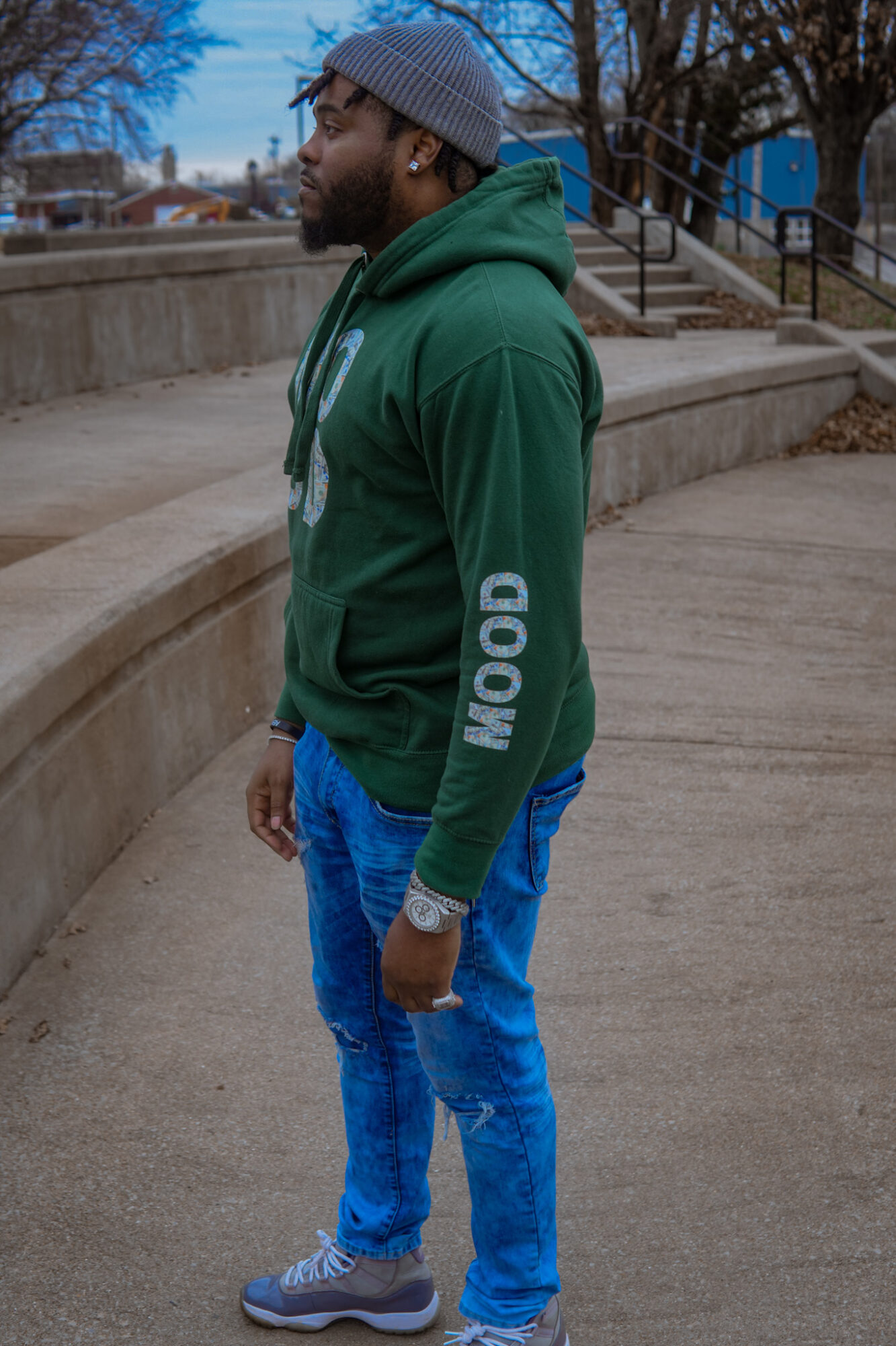 Side view of a man in a green hoodie and blue jeans standing outdoors on a concrete surface.