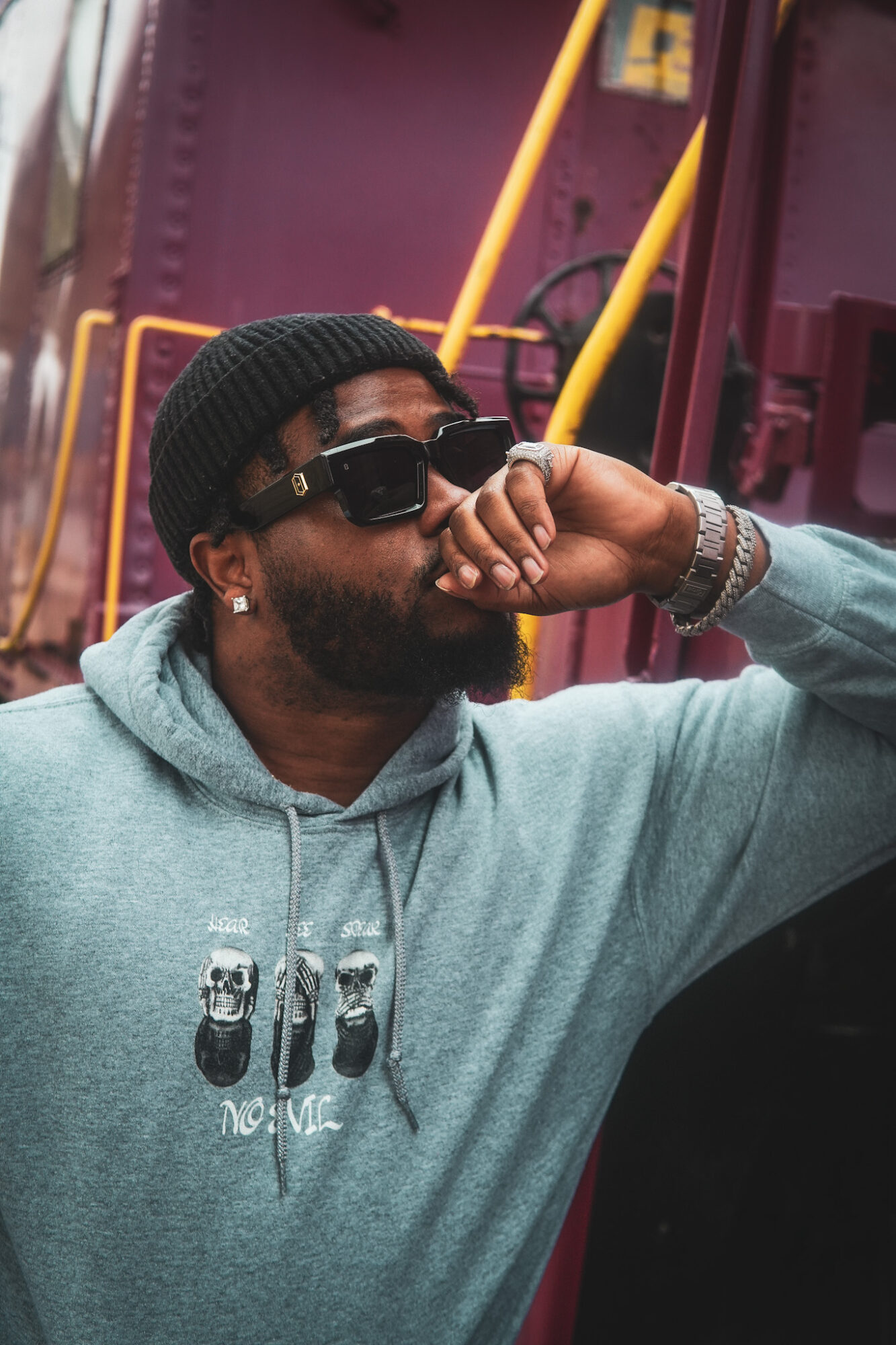 Man with sunglasses and a beanie, wearing a hoodie, sitting with his hand near his face, in front of colorful background.