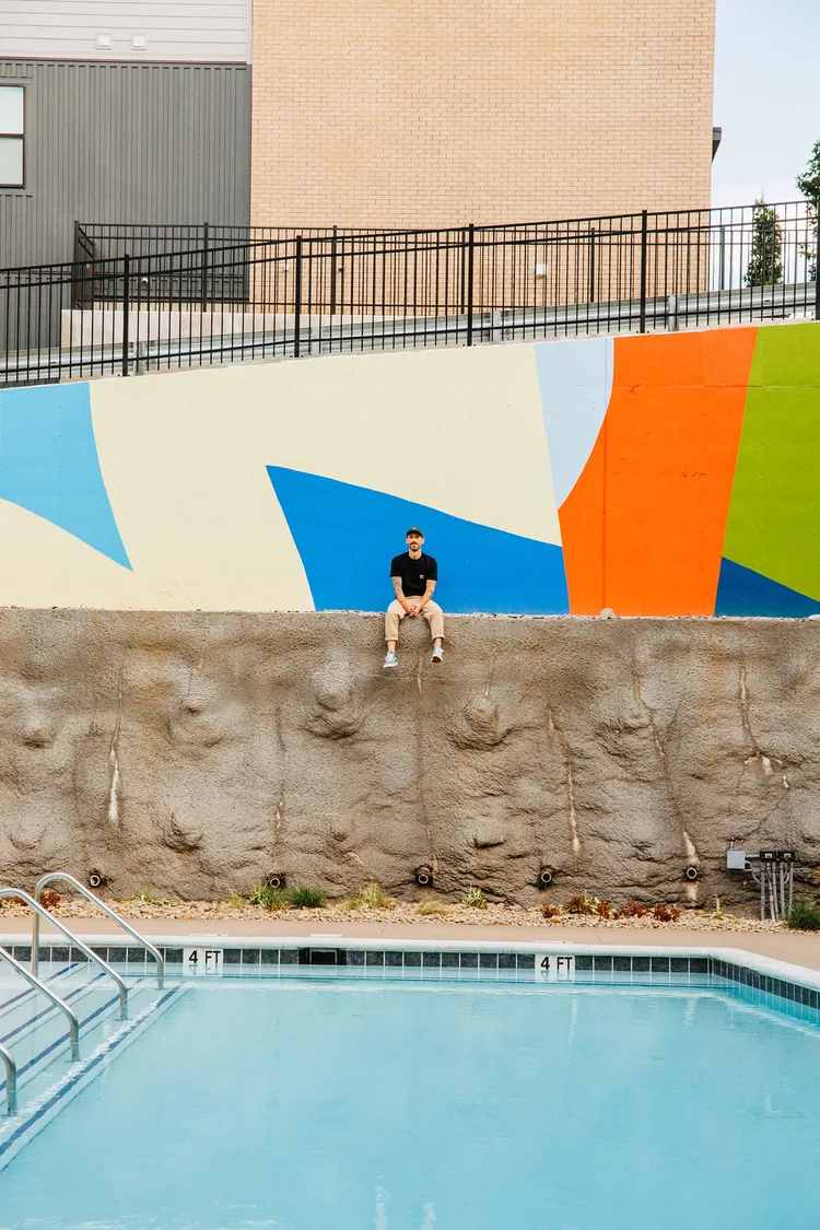 Person sitting on a ledge above a colorful mural near a swimming pool, with a fence and buildings in background.