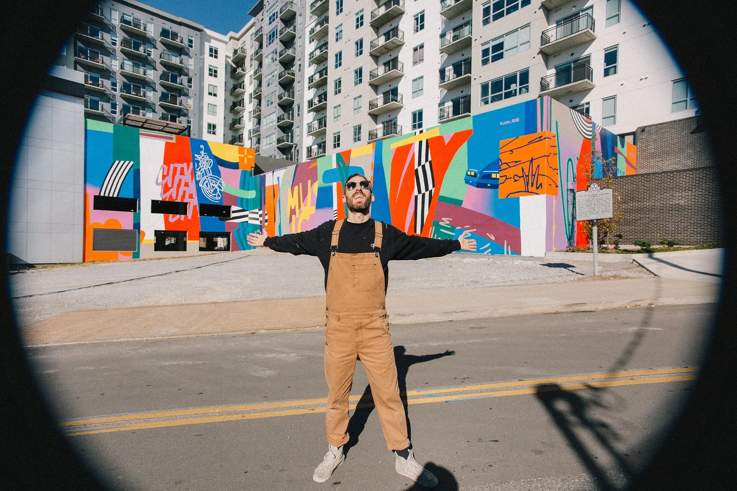 Person standing with arms outstretched in front of colorful mural, urban background with high-rise buildings, sunny day.