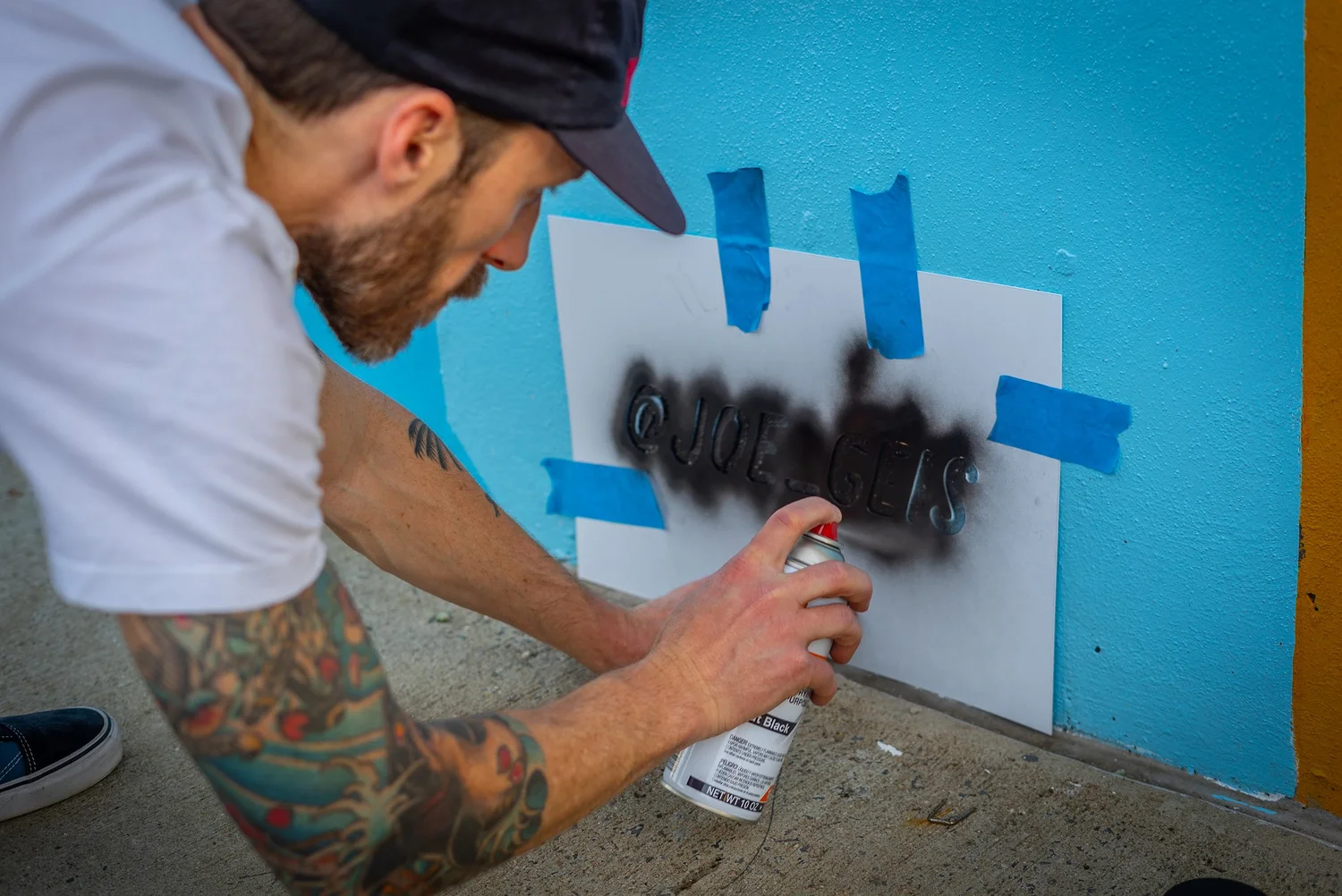Person spray painting graffiti on a white paper taped to a blue wall, with tattoos on arm, wearing a cap.