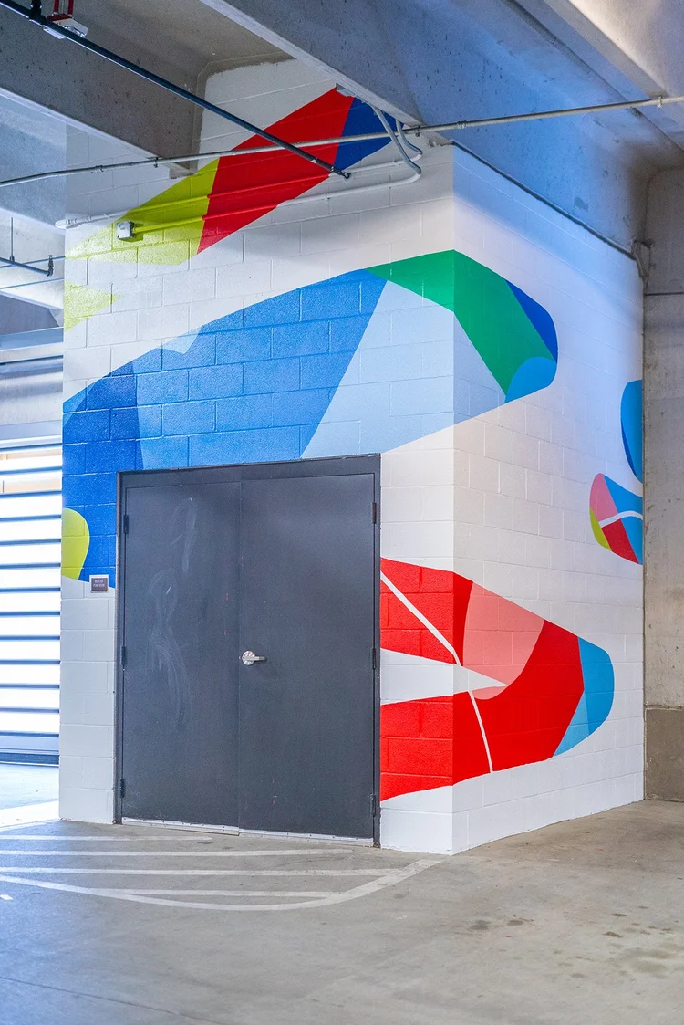 Colorful abstract mural with geometric shapes and patterns on a white wall near a gray door, in an indoor setting.
