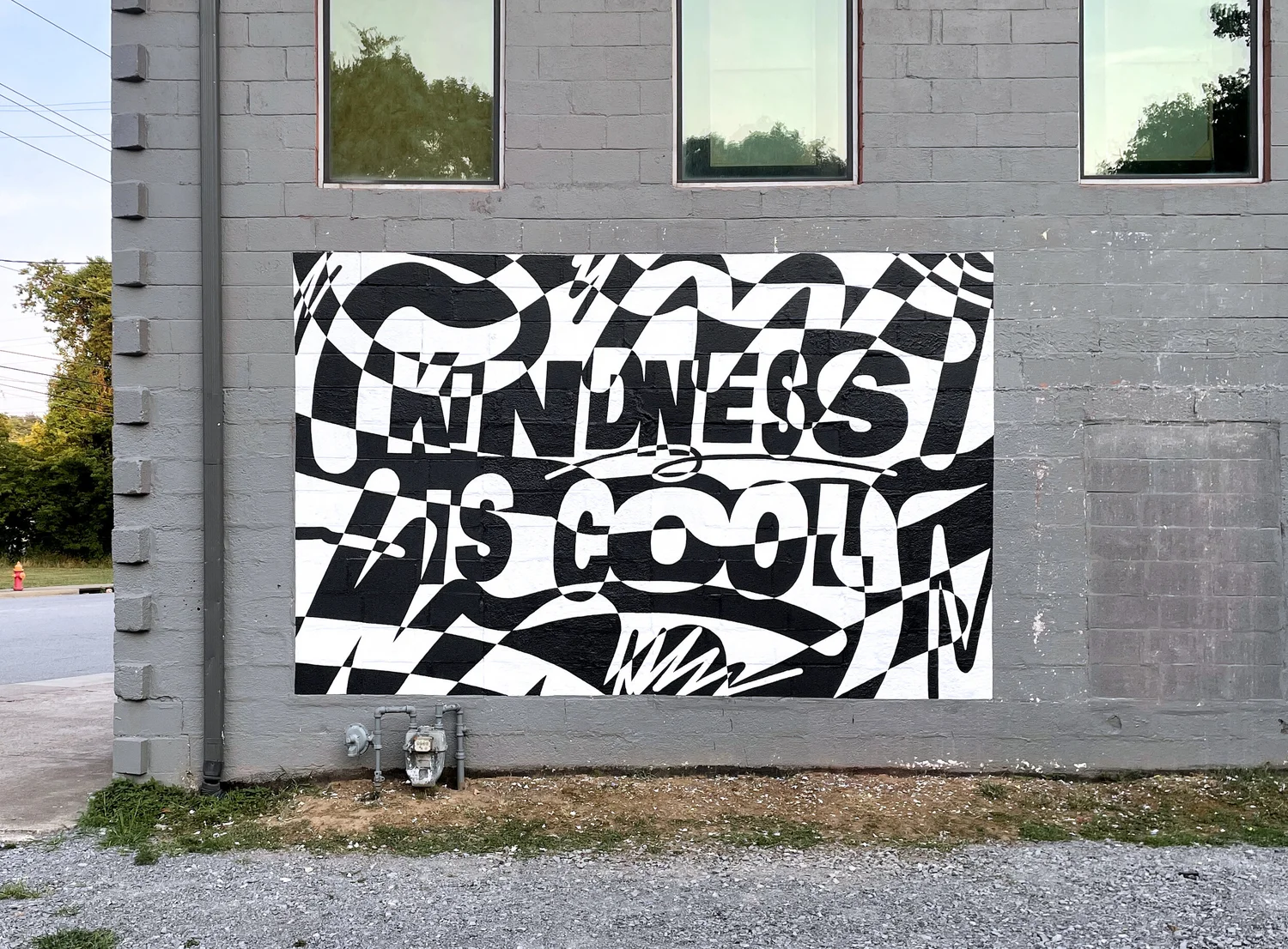 Black and white abstract mural with text reading 'Kindness is cool' on a gray brick wall.