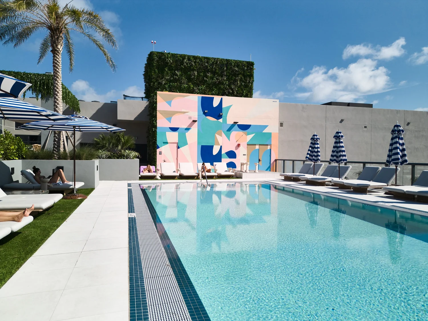 Swimming pool with lounge chairs, umbrellas, and a colorful abstract mural in the background, under a blue sky with clouds.